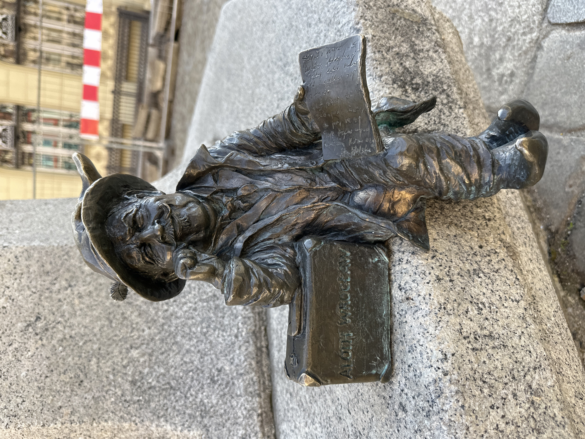 A bronze statue depicting a small, elderly man with a hat, possibly a dwarf, is affixed to a grey stone structure. The man is holding a paper with illegible handwriting, and his other hand rests on a book labeled "ALOHA WROCLAW." The statue appears to be part of a larger installation or memorial. The location is Wrocław, Poland. In the background, a building is partially visible with red and white stripes. The entire scene is shot in daylight.