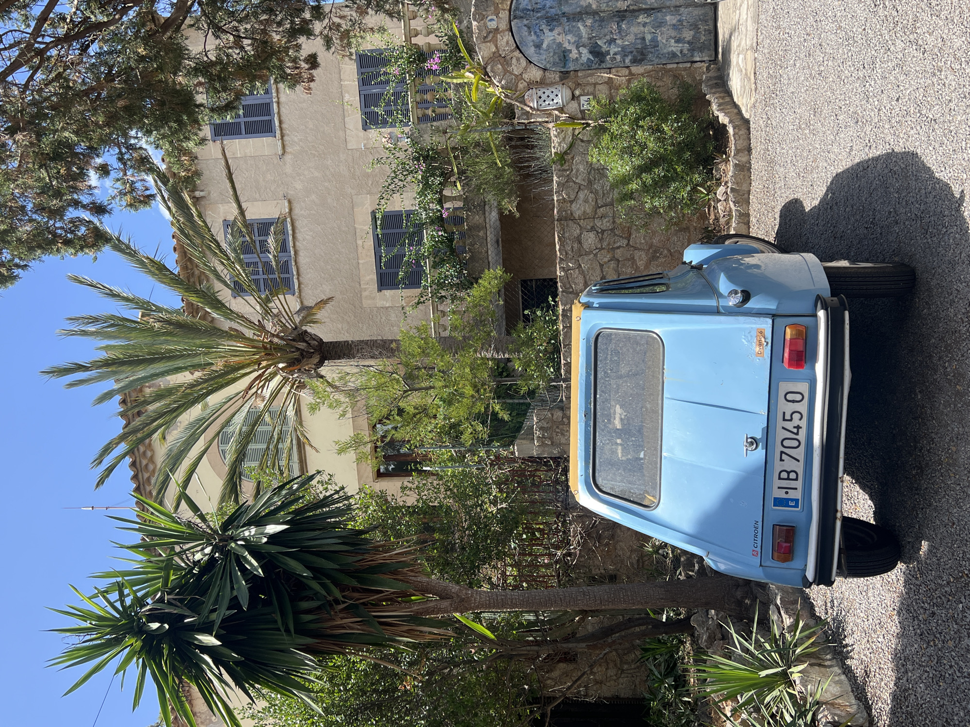 In the city of Pollença, Spain, a classic blue Citroën Dyane 6 is parked on a gravel driveway in front of a two-story tan building with dark blue shutters. A tall palm tree and a yucca plant are visible to the left of the car, with more greenery and trees surrounding the property. The building has a balcony on the second floor, adorned with potted plants. To the right of the building, a rustic stone archway leads to a weathered blue door. The sky is clear and blue. The car's license plate reads "E IB 7045 O".