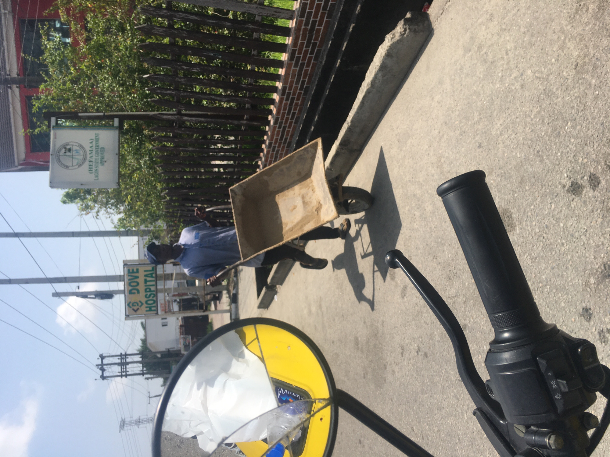 The image, taken from the perspective of someone on a motorcycle, depicts a bright, sunlit outdoor scene, likely on a paved street or pathway. The perspective is slightly angled, showing the motorcycle's black handlebar, grip, and brake lever in the foreground on the right. A yellow-rimmed rearview mirror, cracked and holding some white plastic or paper debris, is prominent on the left.
In the midground, a man is seen pushing a metal wheelbarrow with a tan-colored basin. He wears a light-colored shirt and dark trousers, and his shadow stretches across the paved ground beside him. To the left, a dark wooden fence with a brick base runs along the side of the path, partially obscuring green foliage and trees.
The background features several structures and signs. A white signboard, hanging near the fence, displays a circular logo, the acronym "HEFAMAA", and the text "LAGOS STATE GOVERNMENT APPROVED". Further right, a larger green and white signboard clearly identifies "DOVE HOSPITAL" with a symbol next to the word "DOVE". Overhead, numerous utility poles and a network of power lines crisscross a clear blue sky, dotted with a few white clouds. Portions of buildings, including one with a red roof, are visible behind the signs and foliage.
The scene suggests a functional street environment in a developed area, consistent with the given location context of Ado, Nigeria. The bright sunlight and distinct shadows indicate it is daytime, likely mid-morning or afternoon, under clear weather conditions. The activity of the man pushing a wheelbarrow implies daily work or transport.