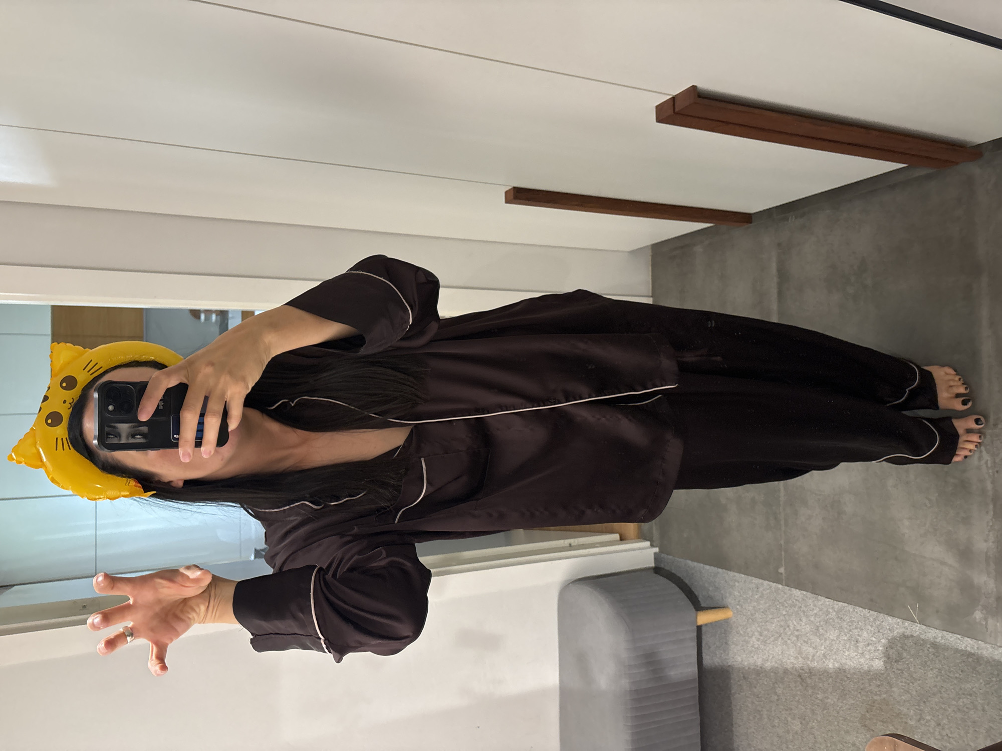 A person stands in a room, taking a mirror selfie. They are wearing dark pajamas with white trim and a yellow cat-ear headband. The person is holding up their left hand with fingers splayed, and a phone in their right hand, obscuring their face. The room has white paneled walls and a concrete floor. To the left, a grey upholstered bench is visible. Two wooden planks lean against the right wall. The scene appears to be indoors, possibly in a home or dressing room. There is no information to indicate this location is in Chaikovsky, Russia.