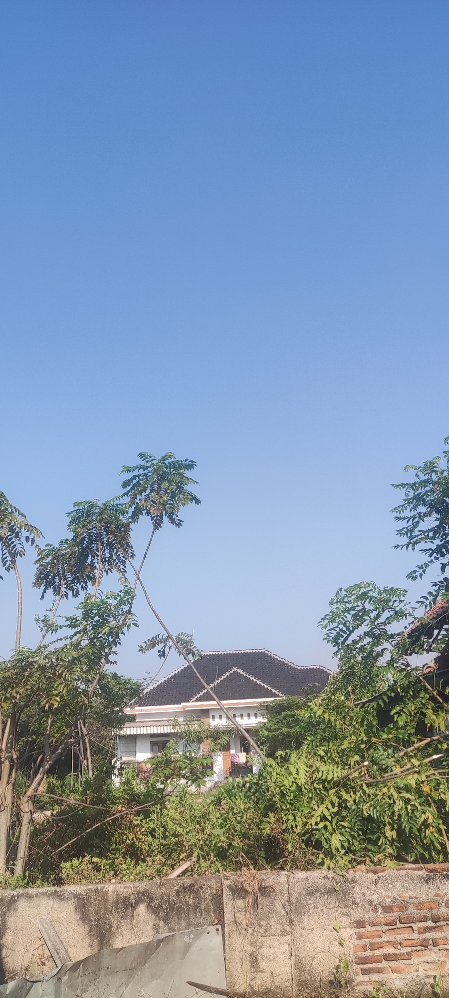 The image depicts a clear blue sky above a residential building in Gebang, Indonesia. The building has a dark tiled roof and white walls with visible windows and decorative elements. Lush green foliage and trees are in the foreground and surrounding the house, obscuring some of its lower sections. A weathered concrete and brick wall is visible in the lower part of the frame, with some discarded metal sheets leaning against it. The scene appears to be captured during daylight, under sunny conditions. No people are visible in the image.