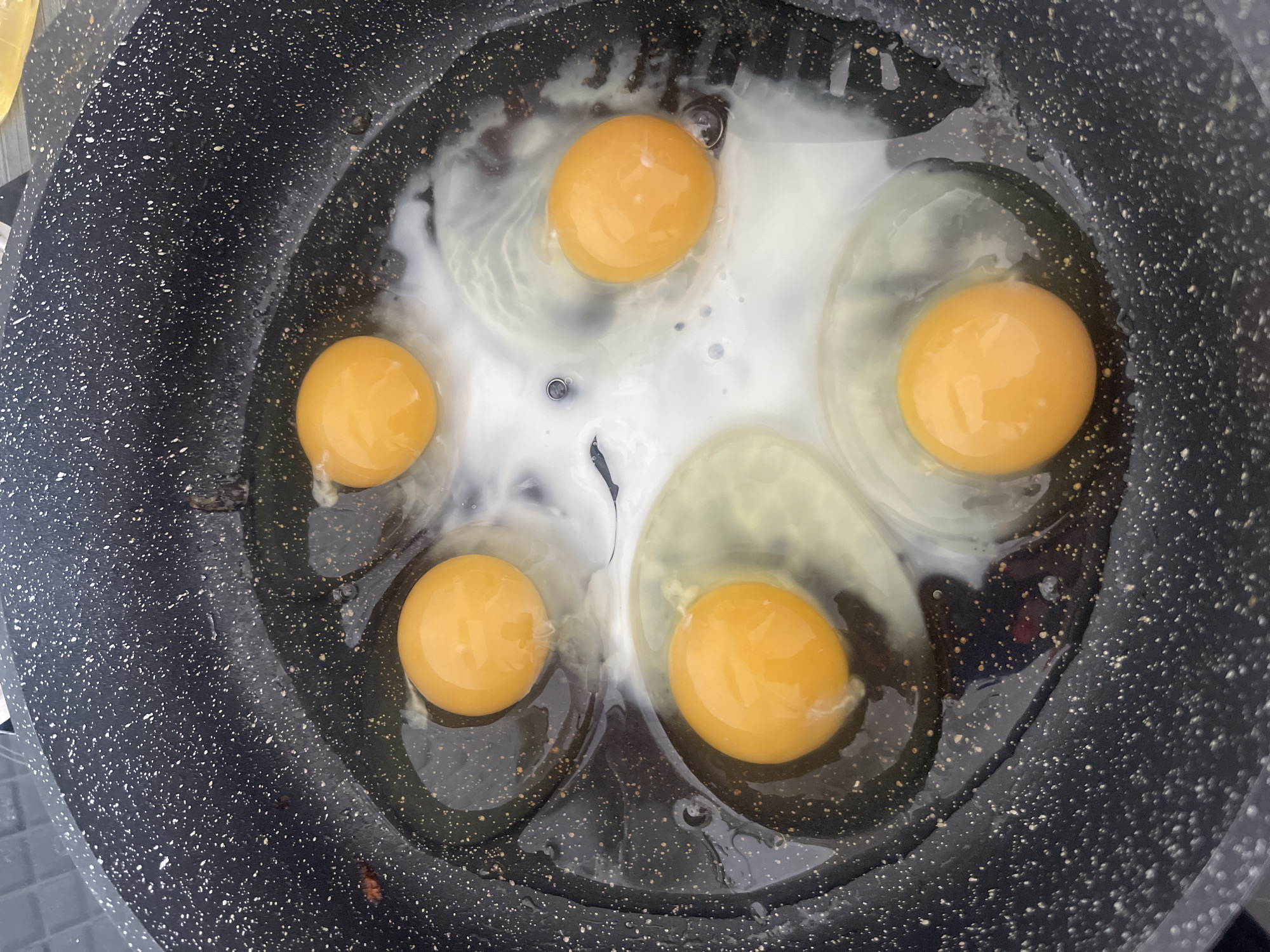 In a speckled black frying pan, five raw eggs are cooking in oil. The yolks are a vibrant orange, and the whites are spread out. The pan is on a dark surface, possibly a countertop, with some light reflecting off it. There is no indication of the location being Blagoveshchensk, Russia.