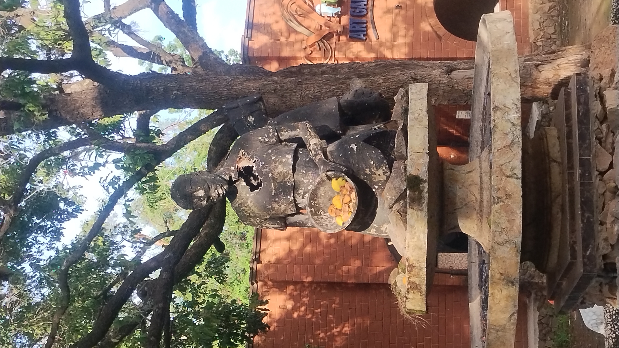 Outside the National Museum in Jos, Nigeria, a damaged statue of a man holding a bowl filled with yellow objects sits partially under a tree. The statue is constructed of a dark material and has a large hole in its chest. Behind the statue, there is a brick building with "Art Gallery" written on its side. There are stone slabs or structures surrounding the statue. The surrounding area is partially shaded by tree branches and leaves.