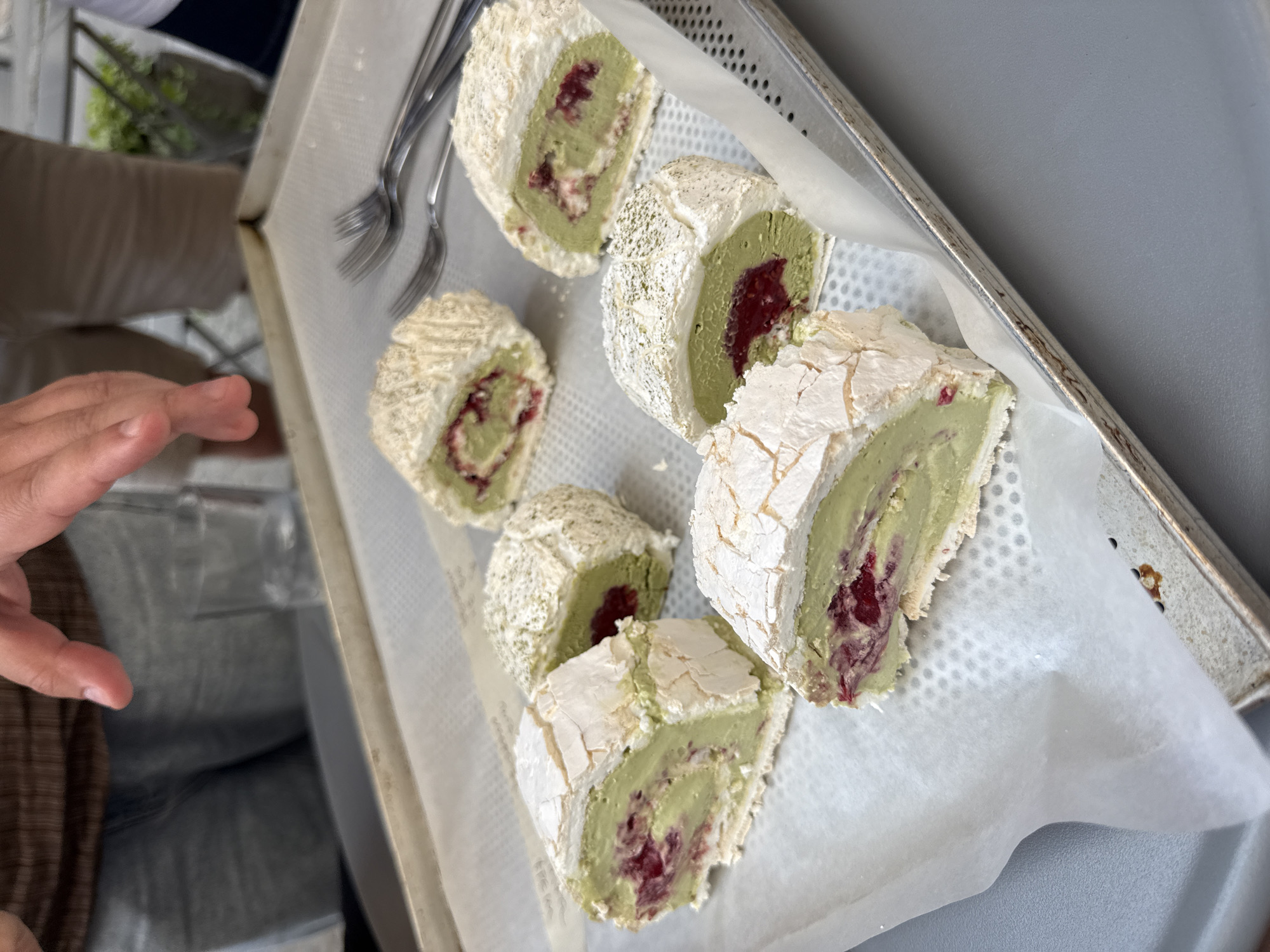 A close-up, slightly angled overhead shot shows six slices of matcha roulade dessert arranged on a silver metal baking tray lined with white parchment paper. The desserts are rolled with a vibrant green filling, possibly matcha cream, and contain a visible red berry component, likely raspberries. The outer layer of each roulade appears to be a meringue, cracked and dusted with a light powder, possibly matcha powder or powdered sugar. Three forks are casually placed on the tray to the upper right of the desserts. In the background, out of focus, are the lower halves of two individuals. One person on the left is wearing a brown patterned shirt and denim jeans, with their hands visible near the tray. The person on the right is wearing tan shorts. The setting appears to be an outdoor cafe or restaurant, suggested by the muted lighting and the presence of what looks like a railing or decorative metalwork in the far background. The overall impression is of a freshly prepared dessert ready to be served. This scene is plausible in Vracar Urban Municipality, Serbia, given the prevalence of cafes and patisseries in the area.