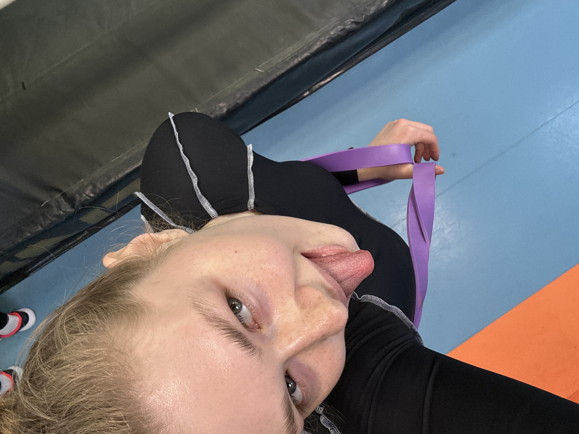 A low-angle, eye-level close-up photo captures a person with fair skin and freckles sticking their tongue out. They have light brown hair. The person is wearing a black, form-fitting garment with white stitching. They are holding a purple exercise band. The background consists of a black surface, a blue surface, and a portion of an orange surface. The location appears to be a gym, possibly in the city of Kazan, Russia.