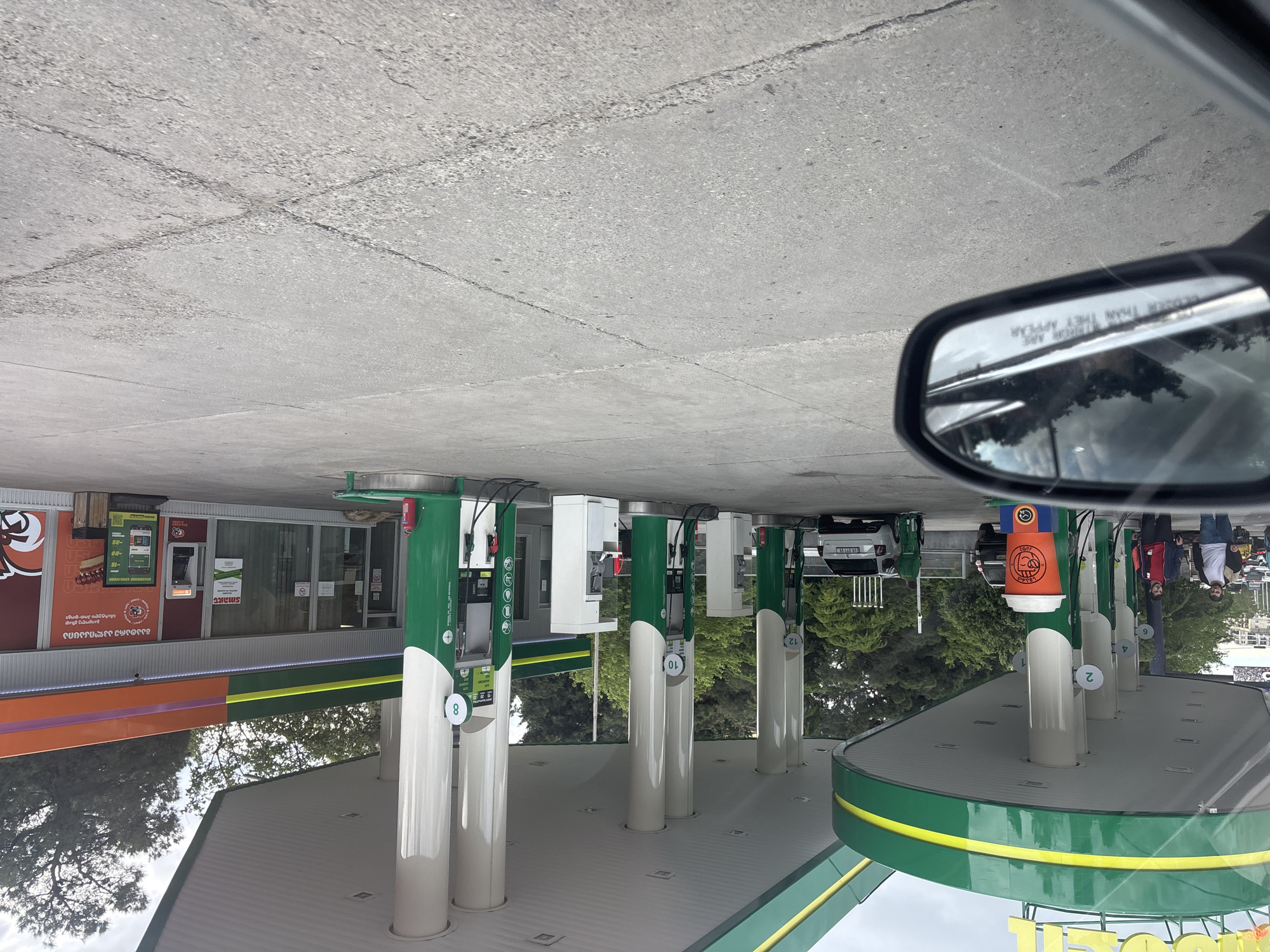 The image depicts a BP gas station in Tbilisi, Georgia. The view is from inside a car, with the side mirror visible in the upper right. Gas pumps are numbered 2, 4, 6, 8, 10, and 12. A sign on the side mirror reads "Objects in mirror are closer than they appear." A white SUV with license plate HVX-011-VX is parked near pump 12. Several people are standing near pumps 2, 4, and 6. A sign for a "smart" hot dog and other food options is visible on the gas station building.
