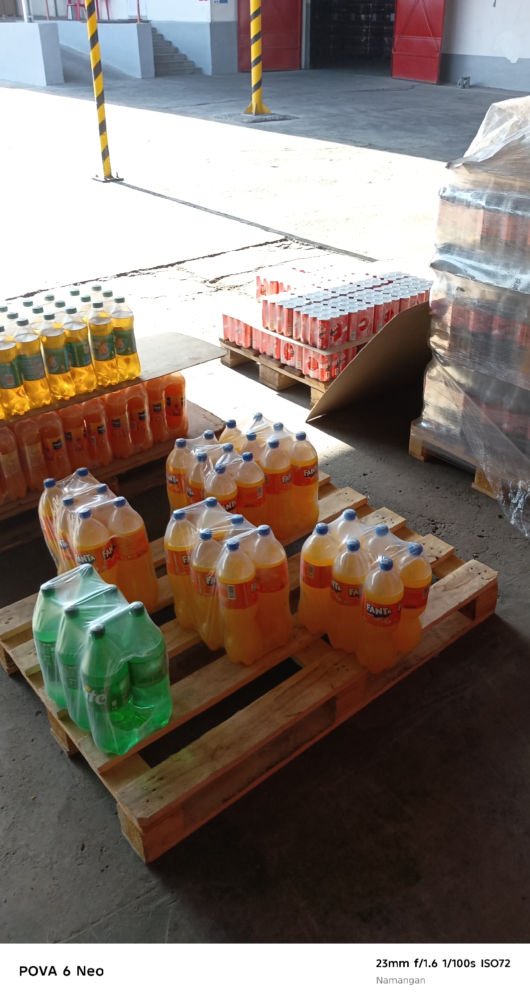 In an outdoor industrial or storage area, likely in Namangan City, Uzbekistan, a collection of beverages is arranged on wooden pallets. The scene is dominated by sunlight casting strong shadows. In the foreground, several rows of plastic bottles containing yellow and green liquids are shrink-wrapped and placed on pallets. Behind these, more bottles of yellow liquid, some in multipacks and others individually, are also on pallets. Further back, stacks of canned beverages, primarily red and white, are visible on pallets, some covered in plastic wrap. In the background, a building with red doors and a grey facade is present, along with two yellow and black striped poles, suggesting a utility area. The ground is concrete, showing some wear and tear. The image appears to be a product shot or inventory photograph of beverages ready for distribution or storage.