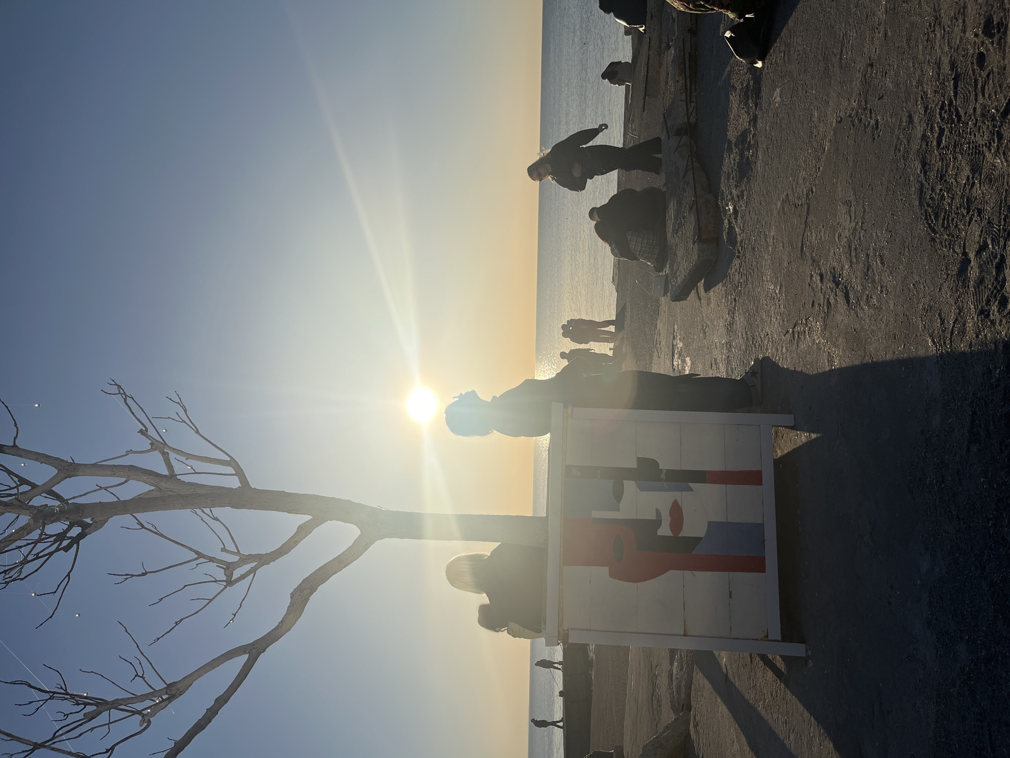 The image captures a sun-drenched coastal scene. Silhouetted figures stand along a breakwater, gazing out towards the sea where the sun reflects. In the foreground, a bare tree stands beside a white sign decorated with a stylized abstract face. The exact location is unknown, but the scene suggests a public space near the coast.