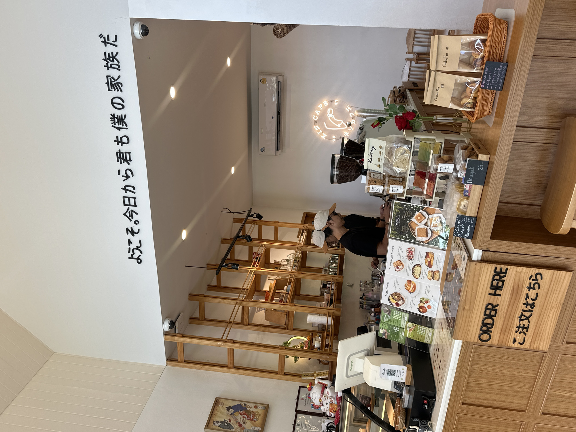 This image depicts the interior of a cafe in Kathu, Thailand. A wooden counter runs across the foreground, with various items for sale displayed on it and behind it. In the center of the counter, a wooden sign reads "ORDER HERE" in English, with Japanese characters below it. To the right of the sign, a woven basket contains bags of baked goods, and a small chalkboard lists "Shoku pan - Cranberry - Chicken floss - Ham cheese." Behind the counter, two baristas, both wearing baseball caps and dark t-shirts, are visible. One is facing the camera, and the other is partially obscured. Shelving units filled with various items, including drinks, food packaging, and decorative elements, are mounted on the back wall. A neon sign that reads "KIN CAFE" in a circular arrangement is illuminated on the back wall, above the counter. The cafe appears to have a modern and minimalist design, with white walls and natural wood accents. An air conditioning unit is mounted high on the back wall.