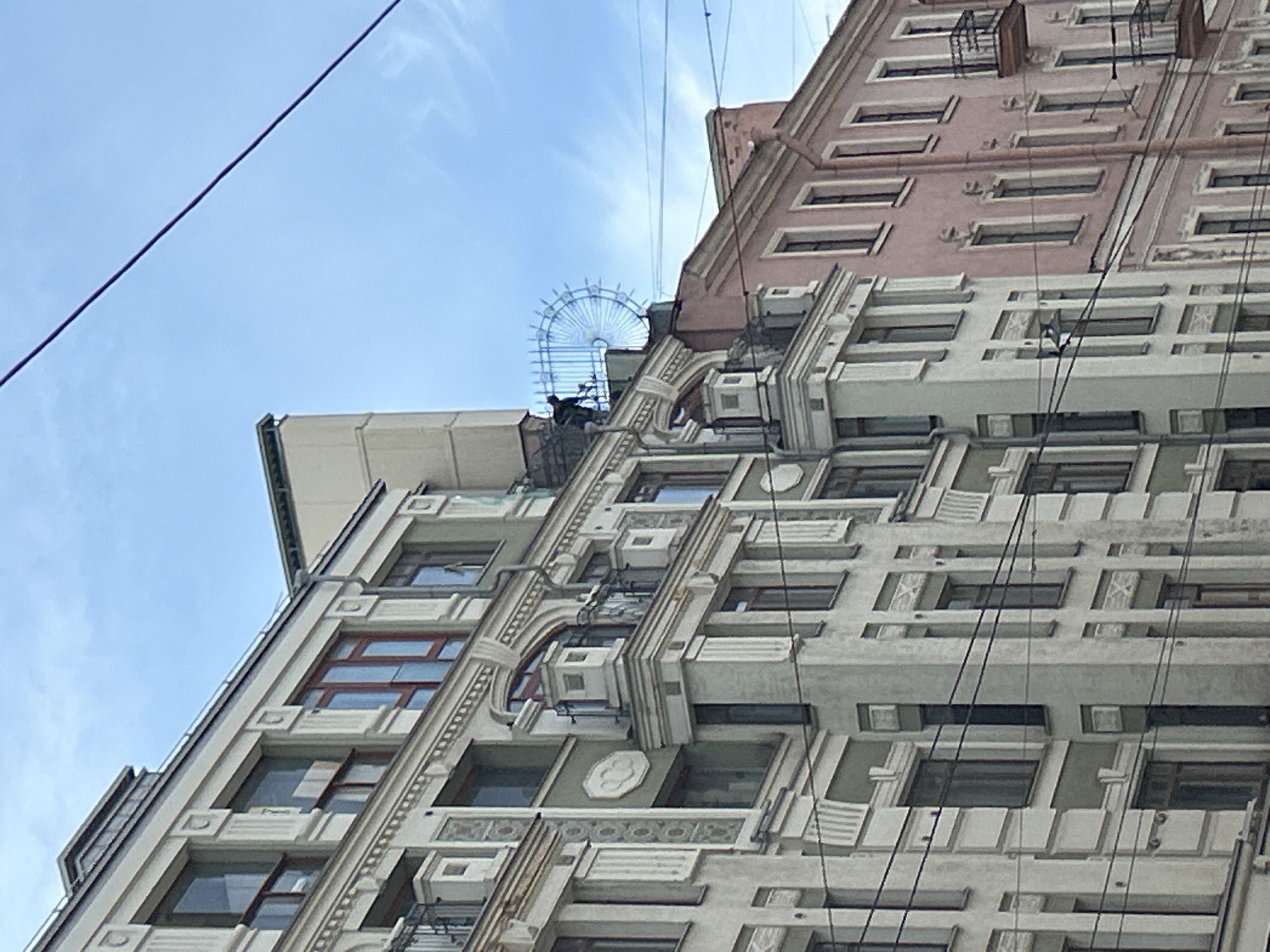 The media file is a low-angle, close-up shot of the facade of a tall, multi-story building against a pale blue sky with wispy clouds. The building has a distinctive architectural style, featuring ornate decorative elements, recessed windows with dark frames, and a layered facade with varying textures and colors, including sections of pale pink and light grey concrete or stucco. Prominent, dark electrical wires crisscross in front of the building, obscuring parts of its details.
On the upper portion of the building, a light blue, semicircular metal structure, resembling a decorative trellis or an incomplete Ferris wheel, is visible against the sky. A dark silhouette of what appears to be a person or a figure is positioned near this structure, possibly interacting with it or observing from above. The overall impression is of an urban environment, possibly historical, with dense wiring suggesting an older infrastructure. The lighting suggests it is daytime, likely late morning or afternoon, given the brightness and the clarity of the sky. There is no visible text in the media file.