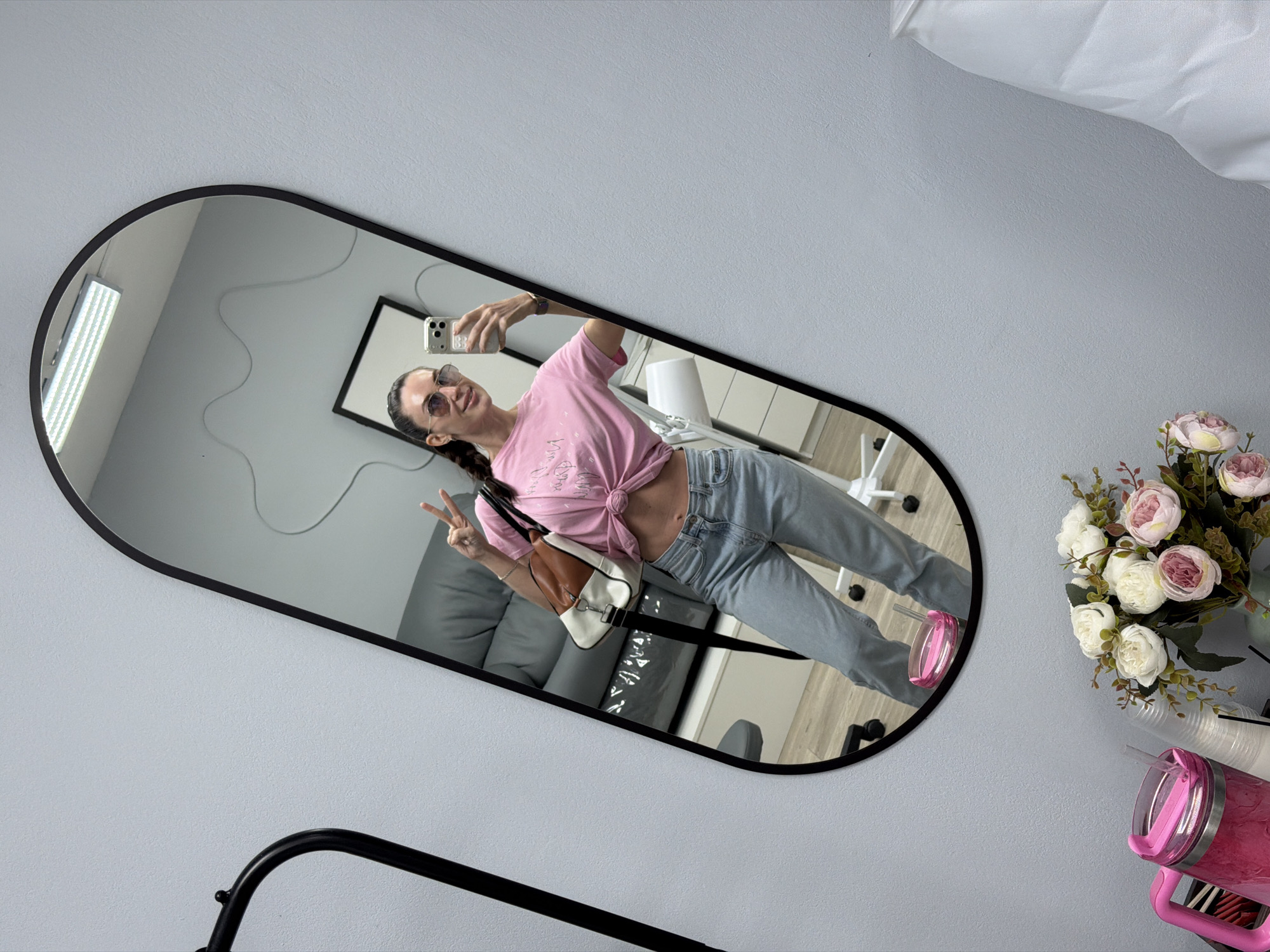 A woman is taking a selfie in a mirror. The mirror is oblong with rounded ends and a black frame and is hanging on a light gray wall. The woman is wearing a pink T-shirt tied in a knot, light blue jeans, and pink shoes. She is also wearing sunglasses and has a white purse on her shoulder. She is holding her phone up with her right hand and making a peace sign with her left. In the reflection of the mirror, a gray couch, a white chair, and a black framed picture on the wall can be seen. To the right of the mirror is a bouquet of pink and white flowers and a pink thermos with a handle. Below the mirror, a black metal rack is visible. The location is the city of Perm, Russia.