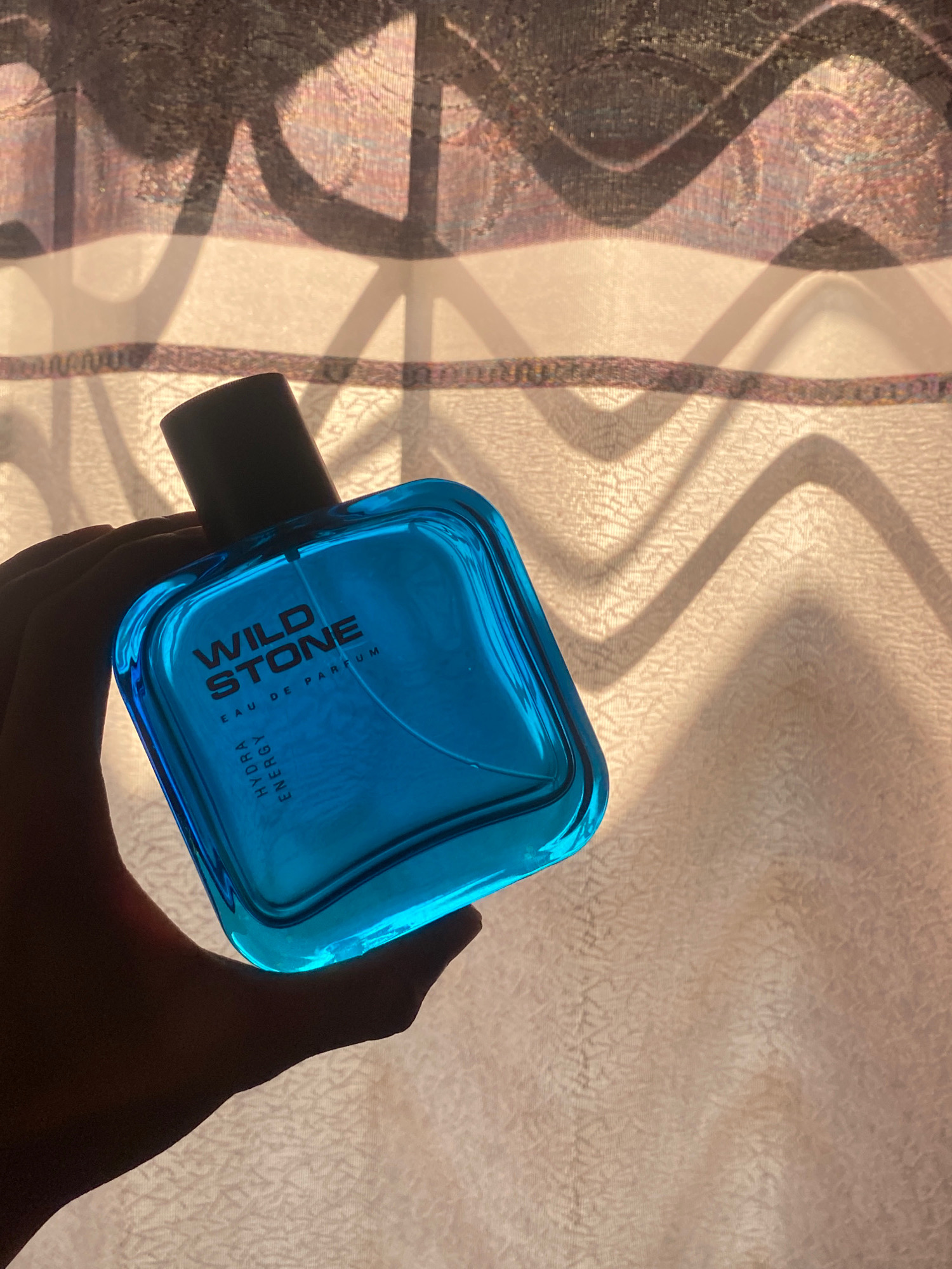 A hand is holding a blue glass bottle of "WILD STONE" eau de parfum, positioned in front of a sheer curtain with a wavy shadow pattern. The bottle is square with rounded edges and a black cap. The brand name "WILD STONE" is printed in red on the bottle, with smaller text below reading "EAU DE PARFUM" and vertically, "HYDRA ENERGY". The background suggests an indoor setting with sunlight filtering through the curtain, casting distinct shadows. The image's composition places the perfume bottle as the central subject. The context of Hemayetpur, Bangladesh, is not visually discernible from the image.