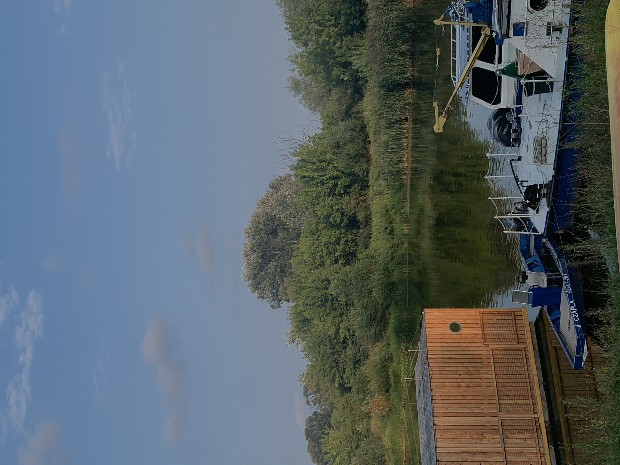 This is a photograph of a tranquil waterside scene in Poznan, Poland. The image is dominated by the calm, reflective surface of a river or lake, with dense green foliage lining the opposite bank, forming a natural backdrop.
In the foreground, to the left, is a modern wooden structure, possibly a house or a boathouse, floating on the water. It has a distinctive circular window and a latticed balcony or railing. Moored alongside this structure are two boats. The smaller, blue boat in the immediate foreground bears the inscription "CZAPLA BG-02-261" on its side. Behind it, a larger, white and blue boat is visible, equipped with a Yamaha outboard motor and a yellow crane-like apparatus.
The sky above is a pale blue, with scattered, wispy clouds adding texture to the upper portion of the frame. The overall impression is one of peacefulness and natural beauty, likely a serene spot on the outskirts of Poznan.
