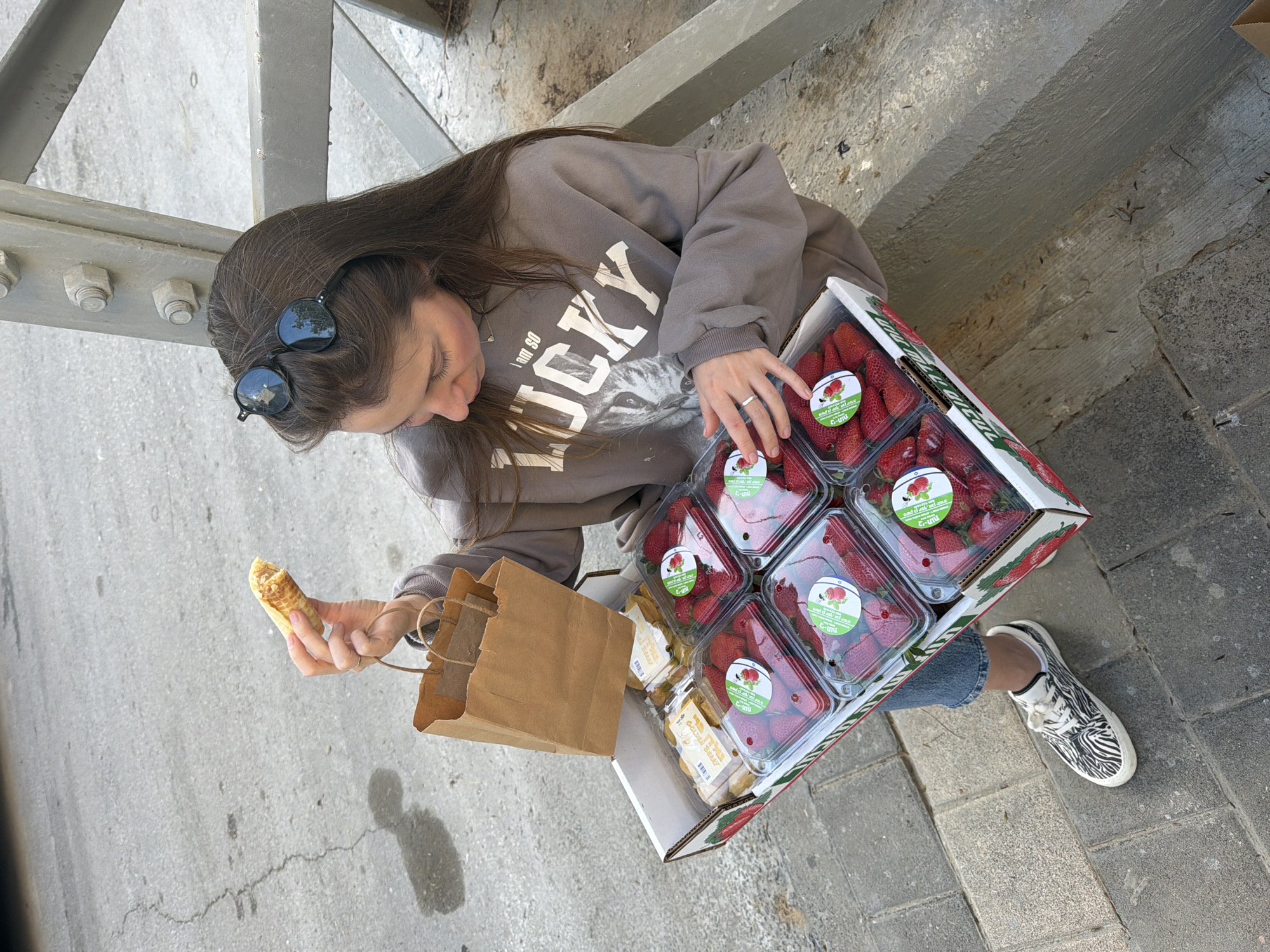 A woman stands on a paved surface in an urban setting, likely the city of Bat Yam, Israel, given the Hebrew writing on the strawberry containers and the fact that Bat Yam is a coastal city. She is holding a filled waffle cone in her left hand and a brown paper bag dangles from her left wrist. With her right hand, she rests on a large cardboard box containing several clear plastic containers of strawberries and some boxes of golden berries. She wears a light brown sweatshirt that reads "I am so Lucky" above a graphic of a cat. She also has blue jeans and sneakers with a zebra-stripe pattern. Sunglasses are perched on her head. She is looking down at the waffle cone. The scene is bathed in natural daylight, with a gray concrete structure visible behind her. The ground is a mix of smooth and textured paving, with a visible crack running across the surface.