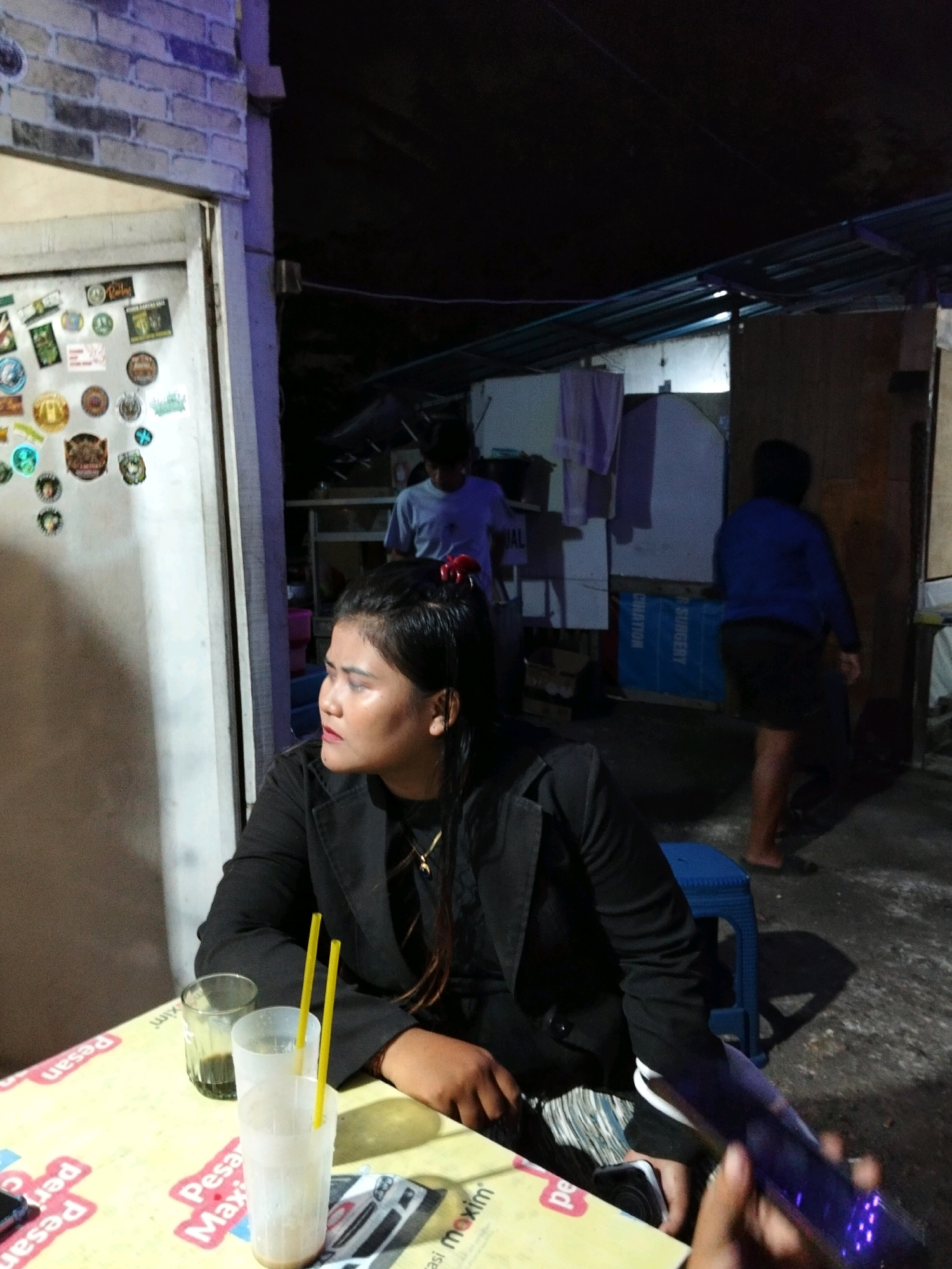 A woman sits at a table with two drinks, one a clear glass with a dark liquid and the other a clear plastic cup with a light-colored beverage and two yellow straws. She wears a black blazer over striped pants and has a red clip in her dark hair. Her gaze is directed to her left. In the background, two other individuals are visible. One person, wearing a light blue t-shirt, stands further back. Another person, wearing a blue long-sleeved shirt and black shorts, is seen walking away. The environment appears to be outdoors, possibly a street-side eatery in Denpasar, Indonesia, with a brick wall and a corrugated metal roof overhead. The table has a yellow patterned surface with some text visible, and a smartphone is on the table.