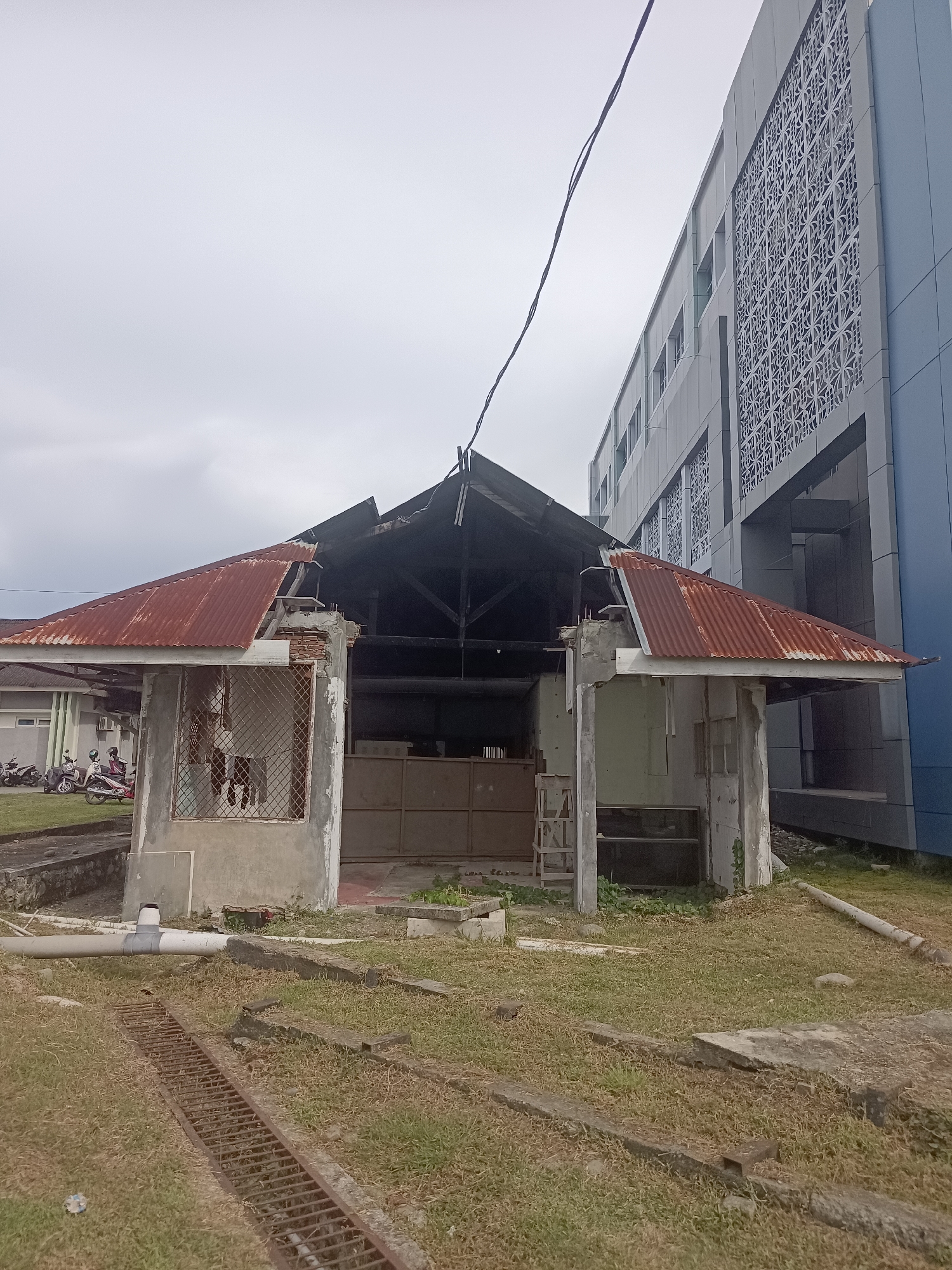 The image depicts an outdoor scene with a damaged, open-air structure in the foreground. The structure has a rusted, corrugated metal roof that is partially collapsed, exposing the wooden beams and framework beneath. The walls appear to be made of concrete with some sections missing, revealing a wire mesh window. Inside the structure, there is a large metal door or gate. To the right of this structure is a modern, multi-story building with a distinctive patterned facade and blue siding. In the background, to the left of the damaged structure, a more conventional building with windows is visible, along with several parked motorcycles.
The ground is covered in dry grass, and a rusty metal grate and concrete elements are present in the foreground. A large pipe runs horizontally across the scene in the lower left. The sky is overcast and grey, suggesting a cloudy day, possibly with diffused light. No people are clearly visible in the immediate vicinity of the damaged structure, though they are present on motorcycles in the background. The overall impression is one of disrepair and abandonment of the smaller structure, contrasting with the intact and modern appearance of the larger building. The location context provided is Mesjid Reuntoh, Indonesia.