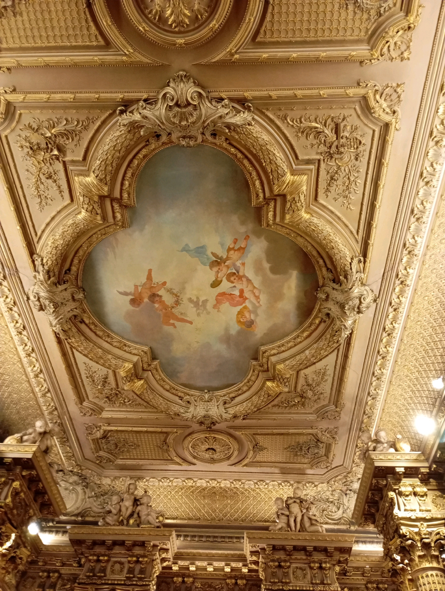 The image shows a highly ornate gilded ceiling, characteristic of Baroque or Rococo architectural styles. The ceiling features intricate relief work with floral motifs, scrolling acanthus leaves, and classical architectural elements. At the center, a painted fresco depicts cherubic figures (putti) flying in a cloudy sky. The painting is framed by elaborate gold stucco or plasterwork. Sculptural elements resembling cherubs are also visible on architectural ledges below the main ceiling. This ceiling is located in the Teatro Colón in Buenos Aires, Argentina, a world-renowned opera house known for its opulent interior design. The lighting appears to be a mix of ambient and artificial light, highlighting the gold accents and the details of the artwork.