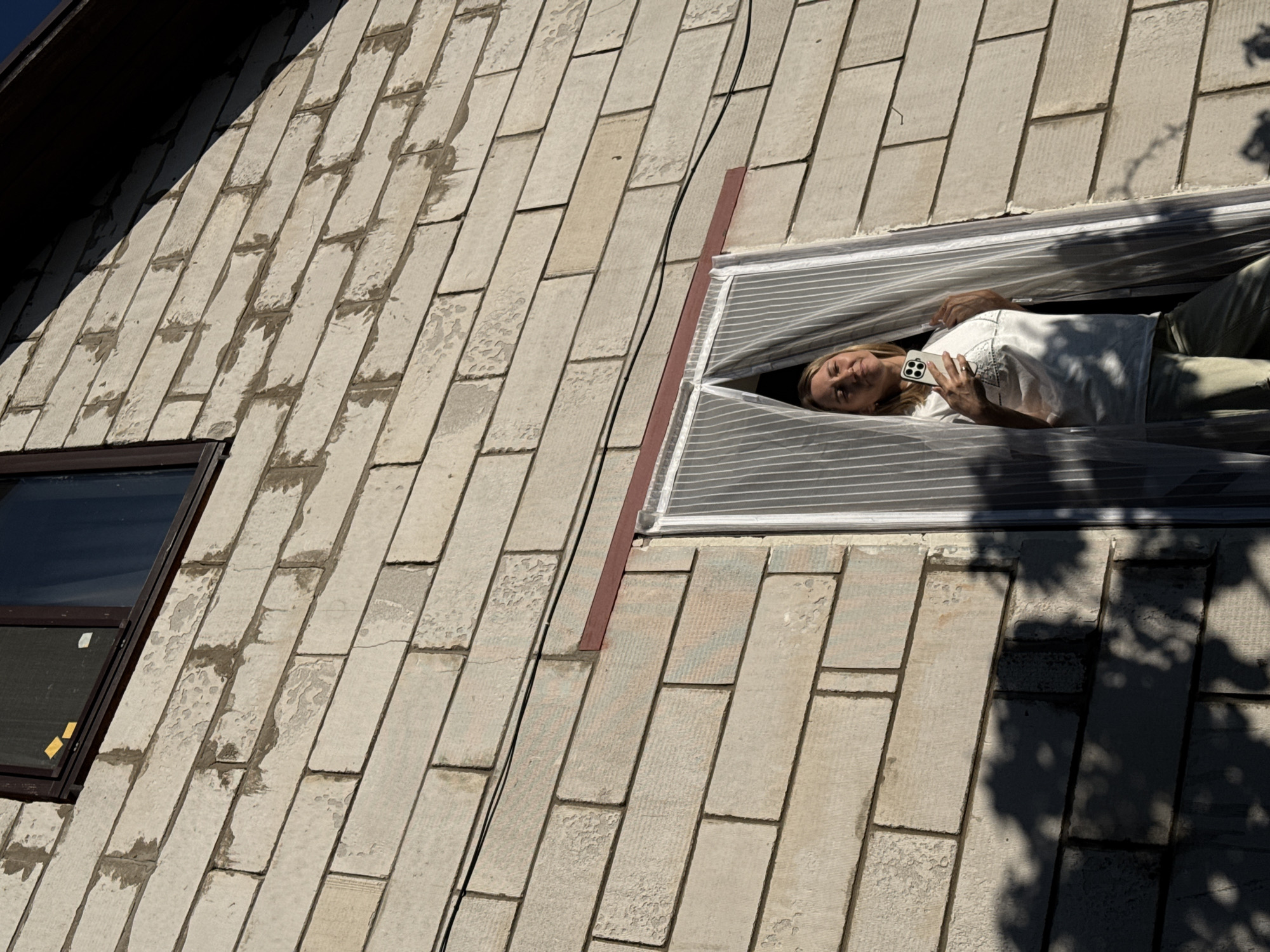 A medium shot depicts the exterior of a light-colored brick building under bright sunlight. A woman with blonde hair is visible through a window that appears to be covered with striped plastic sheeting. She is lying down, holding a white smartphone, and looking towards the camera. There's another window to the left, with its frame in a dark brown color. A dark cable runs from the window across the brick wall. Shadows, possibly from trees, are cast on the brick surface to the right. The image was taken in Chaikovsky, Russia.