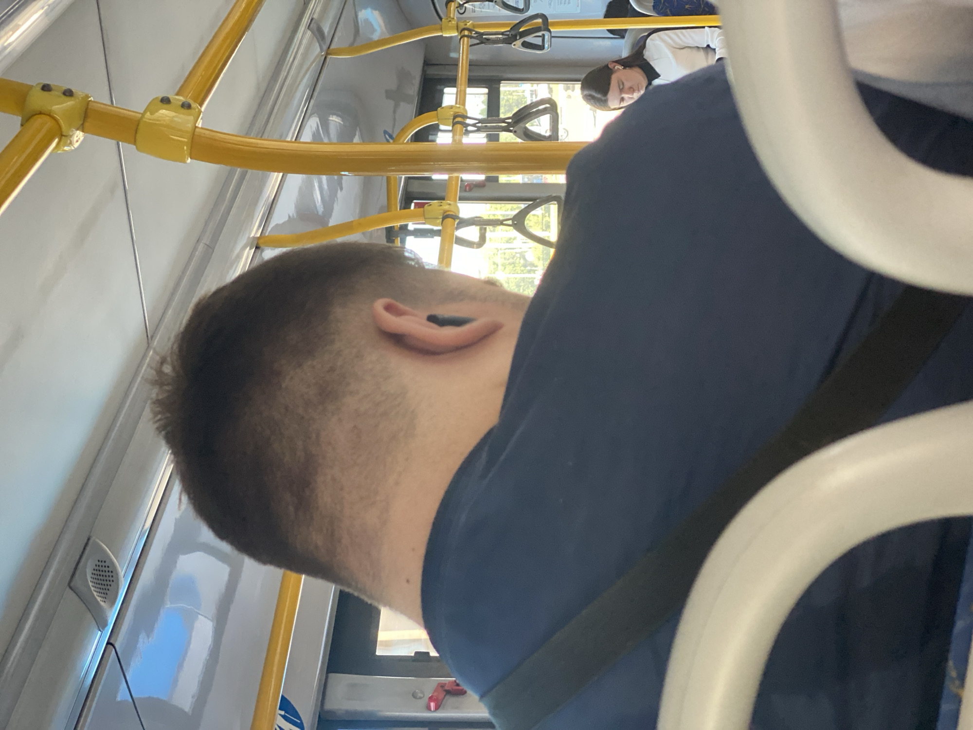 The image shows the interior of a bus in Novokuznetsk, Russia. The viewpoint is from behind a passenger with a dark blue t-shirt and a short haircut. The passenger is wearing black earbuds. In the background, a young woman with long brown hair is seated, also wearing earbuds and a white and black top. The bus has yellow handrails and poles, and white plastic seating. The lighting suggests it is daytime, with sunlight visible through the windows, reflecting on the bus interior. The overall scene depicts a typical public transportation environment.