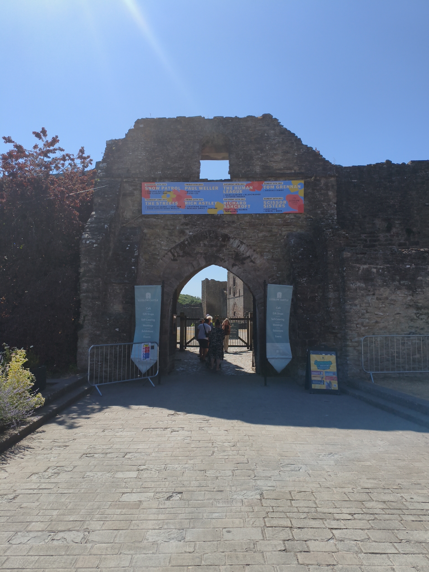 The image depicts the entrance to Ludlow Castle in Ludlow, United Kingdom. The entrance is a large, stone archway, with remnants of the castle walls on either side. A banner is hung across the top of the archway, advertising a series of musical events scheduled for July, featuring artists such as Snow Patrol, Paul Weller, Tom Grennan, and Scissor Sisters.
In front of the archway, people are visible walking through. To the left of the entrance, a lamppost with a blue and white sign for "Ludlow Castle" is present, listing amenities like a cafe, gift shops, and exhibitions. Metal barriers are set up on either side of the pathway leading up to the entrance. A pavement made of stone slabs leads into the castle grounds. The sky is clear and blue, indicating a sunny day. The environment appears to be a historical site with a vibrant event schedule.