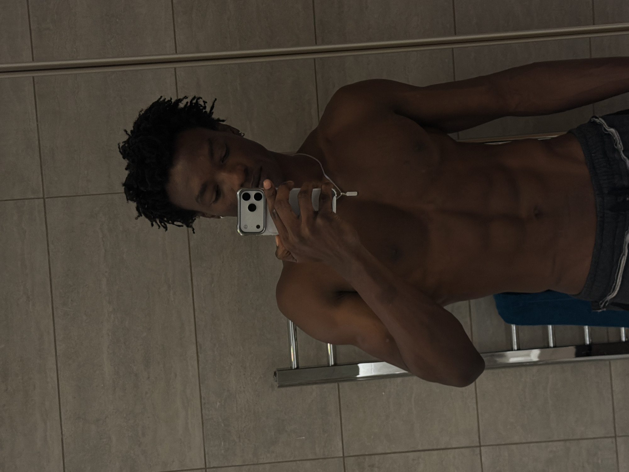 This image, located in London, United Kingdom, depicts a young adult Black male taking a mirror selfie in what appears to be a bathroom. He is shirtless, showcasing a muscular physique with defined abdominal muscles. He wears dark-colored athletic shorts, a silver chain necklace with a small rectangular pendant, and small silver hoop earrings. His dark, curly hair is visible. The man holds a silver smartphone, featuring a multi-lens camera setup, in his right hand, directing it towards his reflection. The background consists of light grey rectangular tiles with horizontal grout lines. A chrome ladder-style heated towel rail, with a blue towel draped over its lower rungs, is visible on the left side of the frame.