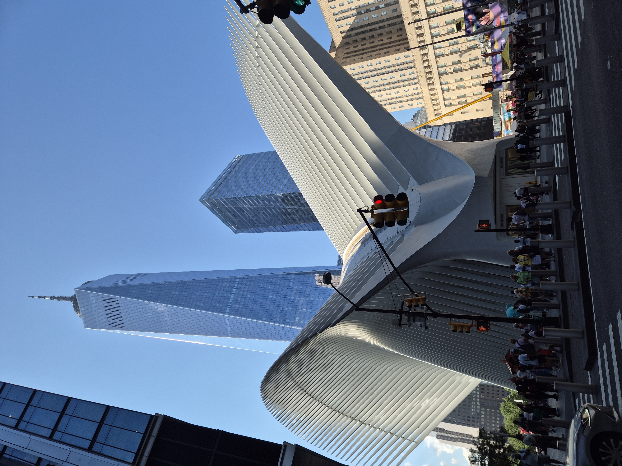 The image captures a street-level view of the Oculus transportation hub, a striking piece of modern architecture located in the Financial District of Lower Manhattan, New York City. Dominating the background is the One World Trade Center, its glass facade reflecting the clear blue sky. To the left, a portion of another modern glass skyscraper is visible.
The Oculus itself is characterized by its white, wing-like structure, composed of numerous ribbed panels. A pedestrian crossing is in the foreground, with a silver car partially visible on the left side. A traffic light with a red signal is illuminated, and a pedestrian signal shows a red walking person. Several people are gathered on the sidewalks and around the Oculus, some walking and others standing. Behind the Oculus, a tall, beige building with numerous windows is visible, alongside other urban structures. A yellow construction crane extends upwards behind the Oculus. The overall scene is bathed in bright sunlight, casting shadows and highlighting the architectural elements.