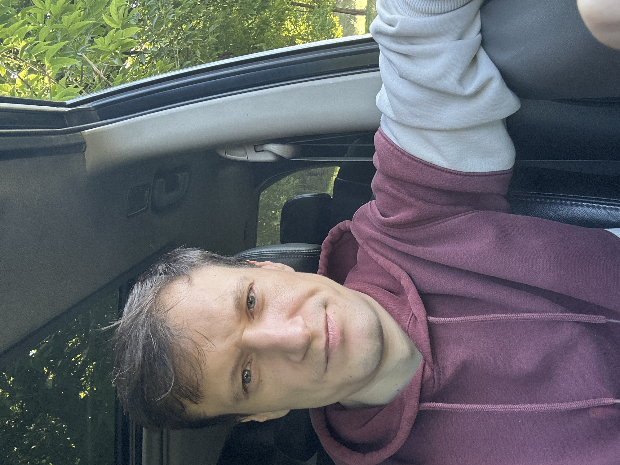 A man is seated in the driver's seat of a vehicle, possibly an SUV given the view through the sunroof and the side window. He is looking directly at the camera, with a neutral expression. He is wearing a maroon hoodie with contrasting cream-colored sleeves. The interior of the car appears to be dark, with black leather seats visible. Outside the car, green trees and foliage are visible, suggesting an outdoor setting, possibly a park or a rural area. The lighting indicates it is daytime, with sunlight filtering through the leaves. The photo is taken from a slightly low angle, looking up at the man's face.