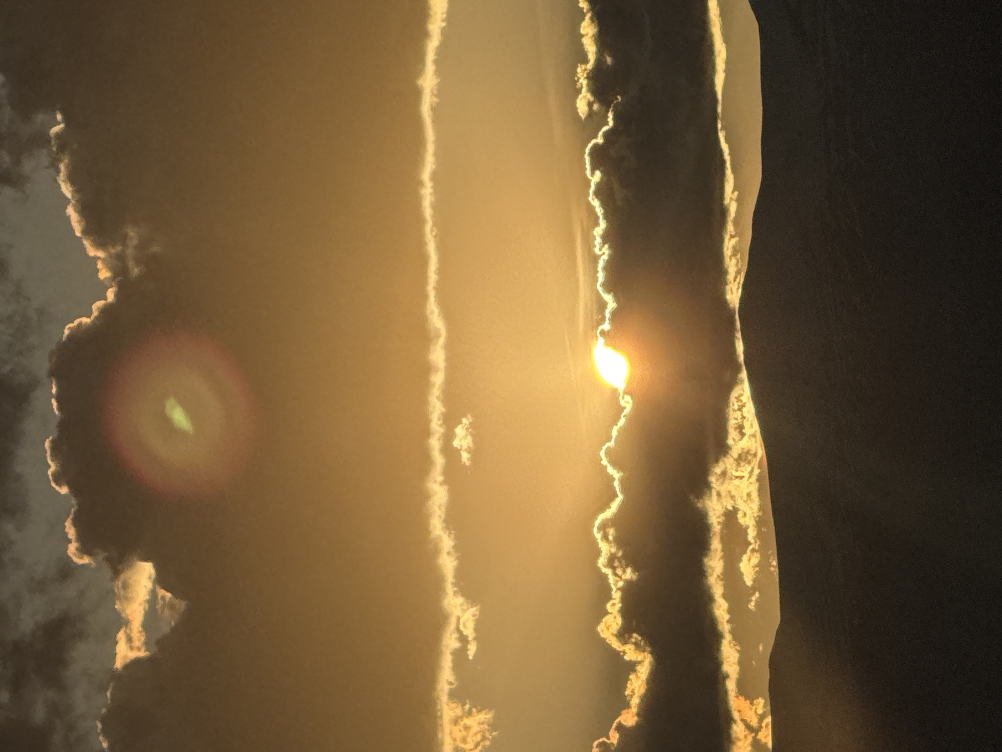 The image displays a dramatic and vibrant sky scene, likely capturing either a sunrise or sunset. The photograph is oriented sideways, with the sun positioned prominently on the right side of the frame, partially obscured by dense cloud formations.
The primary subjects are the sky, clouds, and the sun. The sun's intense light, a brilliant yellow-white, peeks from behind a jagged cloud edge, illuminating the surrounding clouds with fiery golden and orange hues. These backlit clouds form distinct, somewhat vertical bands stretching across the frame, with their sun-facing edges glowing brightly against the darker, shadowed masses. Unlit sections of the clouds appear in deep gray and black, especially towards the far right edge of the image.
A notable feature is a prominent circular lens flare located on the left side of the image, characterized by a luminous green center surrounded by concentric reddish-orange rings, set against a darker cloud area. The overall atmosphere is warm and hazy, with the golden and amber light diffusing across the sky, creating a sense of depth and intensity. The scene suggests a specific time of day—either dawn or dusk—due to the dramatic interplay of light and shadow, and the rich color palette. No specific location cues are visible within the image itself, though the context indicates Abovyan, Armenia. There is no visible text in the image.