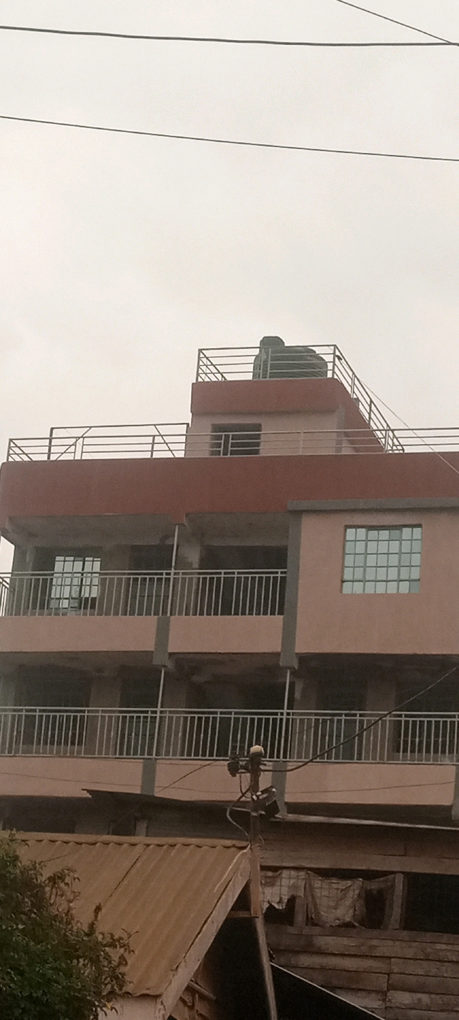 This image, taken in Kabete ward, Kenya, shows the exterior of a multi-story building under construction or recently completed. The building features a reddish-brown facade and balconies with silver railings on at least three levels. There are multiple windows with a grid pattern, some reflecting the overcast sky. On the rooftop, a large dark water tank is visible, along with more railings and what appears to be a small structure or ventilation unit. In the foreground, a corrugated metal roof of a lower structure partially obstructs the view of the building's base. A utility pole with wires and electrical components is also present, situated between the two structures. The overall environment appears to be urban or semi-urban, with a focus on residential or commercial development.
