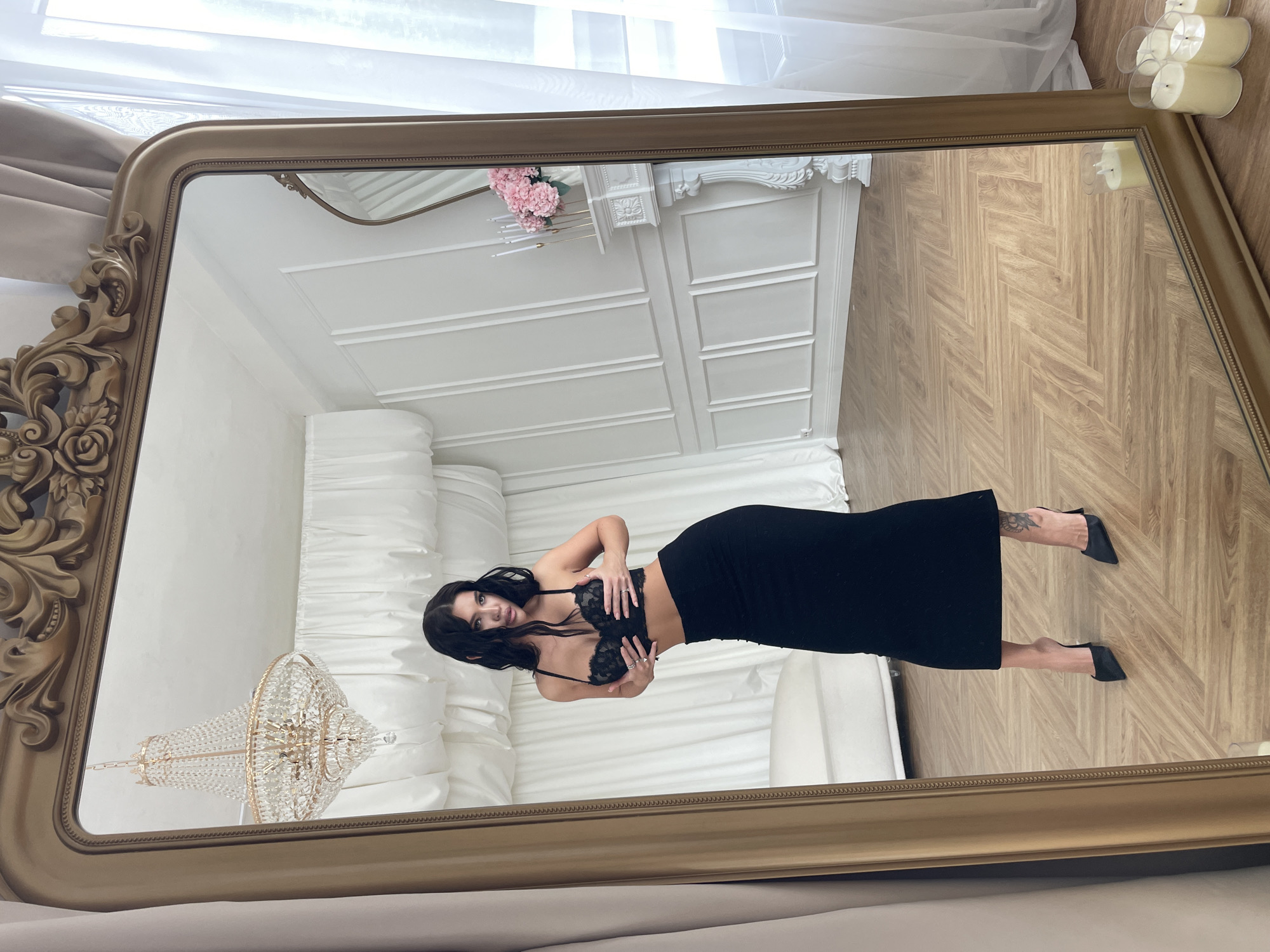 A woman with dark hair, wearing a black lace bra and a black skirt, stands reflected in a large, ornate gold-framed mirror. The reflection shows a lavish interior, featuring a white tufted couch, a crystal chandelier, a white fireplace mantel adorned with pink flowers, and herringbone-patterned wood flooring. Candles are present in the top right corner of the frame. The woman's posture is expressive, with her hands positioned near her chest. The image is well-lit, highlighting the details of the room and the woman's attire. The location in the city of Kazan, Russia is unknown.