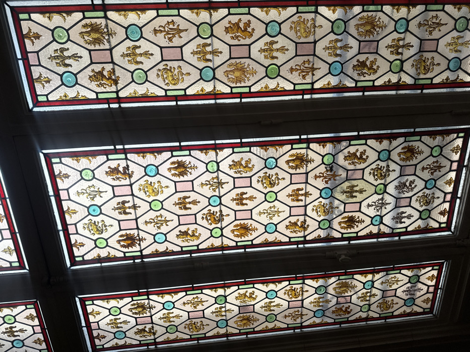 The image displays a series of vertical stained glass window panels, seen from an interior perspective. Four distinct panels are visible, with a partial view of a fifth at the top left. Each panel features a repeating geometric and heraldic design. The primary pattern consists of interconnected hexagonal and square segments, outlined in dark leading. Within these segments, various golden-yellow heraldic motifs are depicted, including fleur-de-lis, rampant lions, eagles, and coiled serpents/dragons, set against backgrounds of clear, pale yellow, or pale pink glass. Circular panes of turquoise and light green glass are interspersed throughout the grid, often at intersections or as central elements within specific hexagonal sections. Each window panel is bordered by strips of red and light green/clear glass. The windows are illuminated from behind by daylight, highlighting the colors and translucency of the glass. The dark framing elements suggest a substantial architectural setting. This specific style and array of heraldic symbols are characteristic of the stained glass located within the Hôtel de Cluny (Musée de Cluny - National Museum of the Middle Ages) in Paris, France. No people or specific actions are depicted.