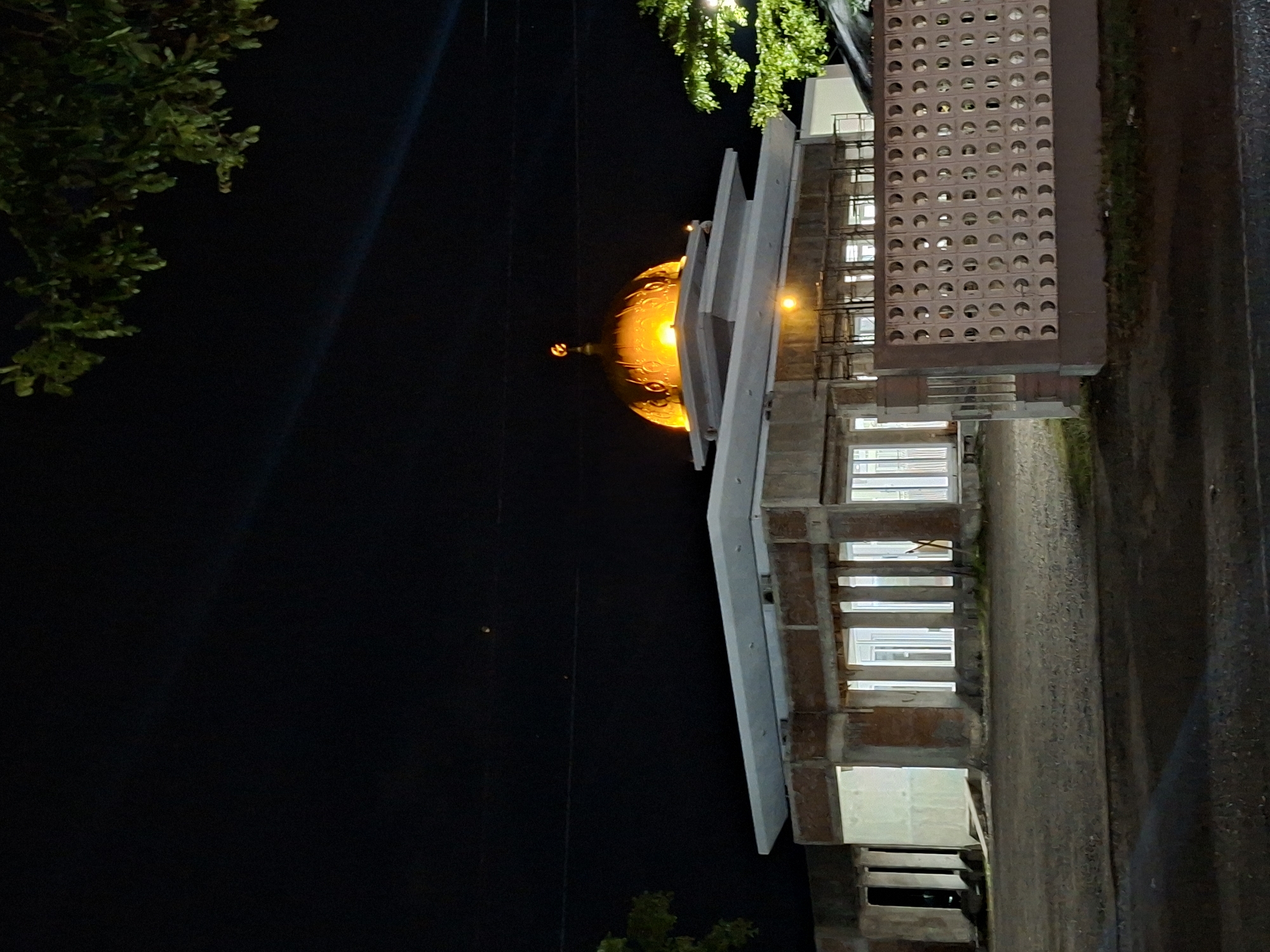 The image captures a nighttime view of a mosque in Reuhattuha, Indonesia, which appears to be under construction or renovation. The central feature is a prominent, brightly illuminated golden dome, crowned with a distinct crescent moon and star finial. Beneath the dome, the building exhibits an unfinished state with exposed concrete beams and pillars, and partial brickwork. The upper roof sections are flat and light-colored, presenting a tiered appearance. The lower section of the building displays multiple vertically oriented white-framed windows, through which interior lighting is visible. Scaffolding-like structures are present on the upper left portion of the building.
The foreground consists of a dark, unpaved ground surface, possibly gravel. To the right, a low, light-colored perimeter wall composed of patterned breeze blocks with circular perforations extends into the frame. Minimal sparse green vegetation is visible at the base of this wall. The sky is dark, indicative of nighttime, and contains faint diagonal light streaks in the upper sections, along with two small, indistinct points of light. Dark green foliage from trees or shrubs partially obscures the view from the upper left and upper right corners of the frame. No individuals or animals are present within the captured scene.