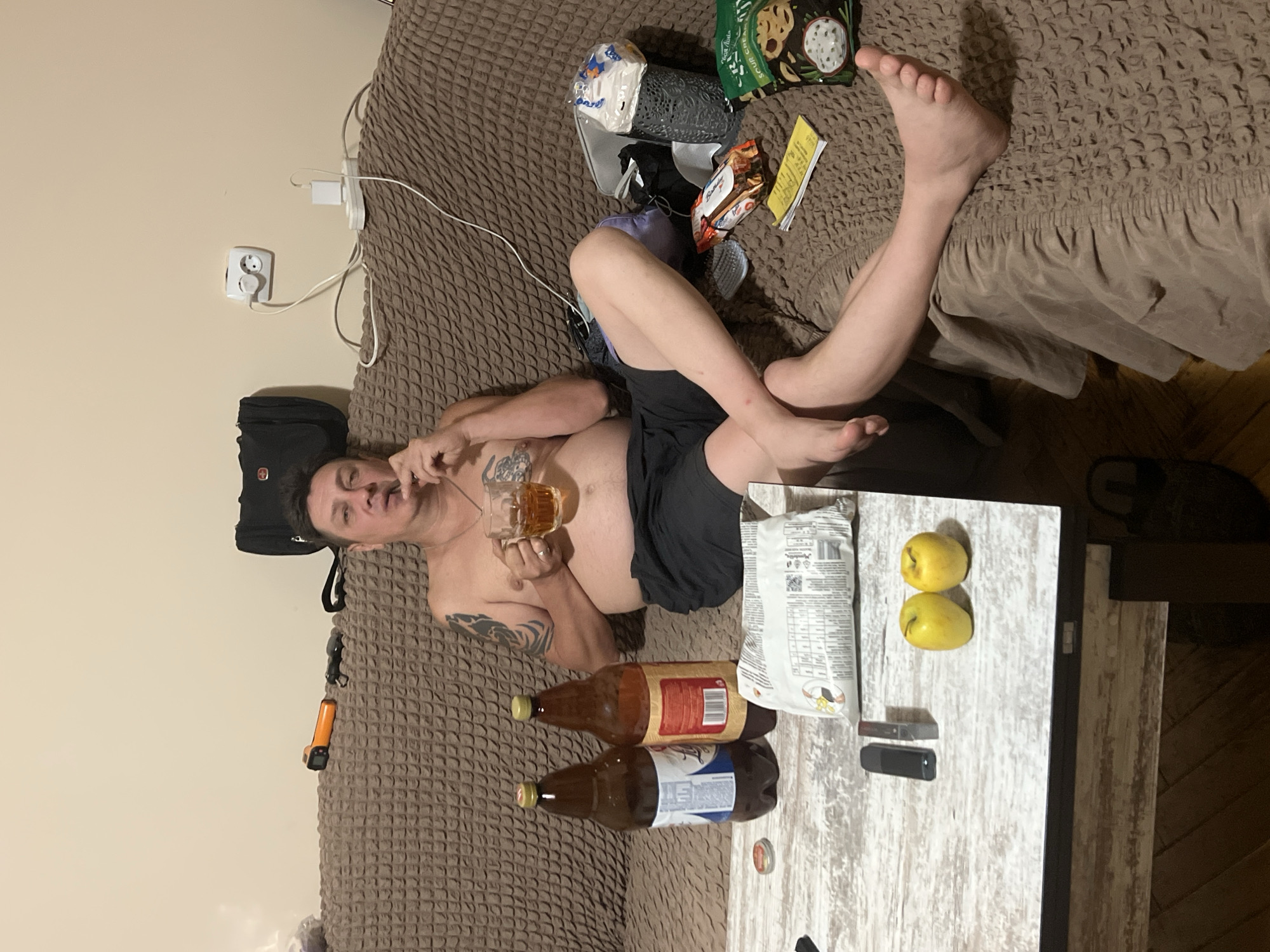 In the city of Drohobych, Ukraine, a shirtless man with tattoos on his left arm is seated on a brown textured sofa, holding a glass of amber liquid. He is wearing black shorts and appears to be smoking. In front of him is a white wooden coffee table. On the table are two large brown bottles, a bag of snacks, two yellow apples, and two dark rectangular objects, possibly electronic devices. The sofa has a brown patterned cover. Behind the man, on the wall, is an electrical outlet with a white power strip and several plugs. A black bag is also visible on the sofa. To the right of the man, on the sofa, are more snacks and a grey textured container. The overall environment suggests an indoor domestic setting.