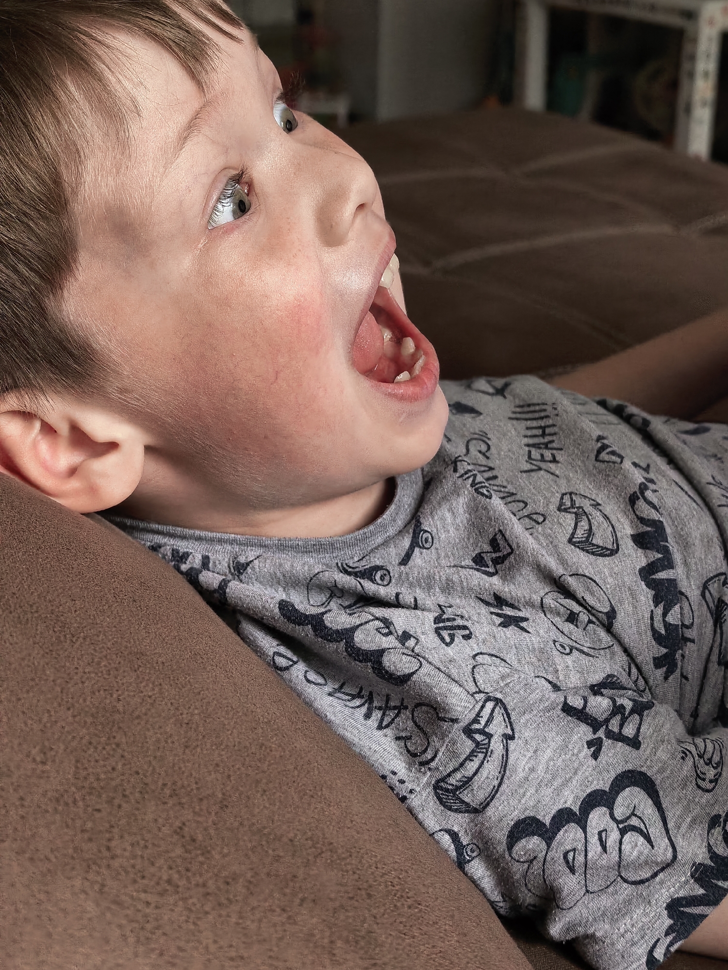 The image captures a close-up of a young child, likely a boy, positioned on a brown upholstered sofa. He is seen from a high angle, lying down with his head slightly raised, looking intensely upwards and to the right of the frame. His mouth is wide open, revealing his lower teeth and gums, indicating a strong expression of surprise, excitement, laughter, or vocalization. His eyes are wide and focused upwards, conveying a sense of wonder or keen interest.
The child has light brown hair and fair skin, with a noticeable flush on his cheeks. He wears a grey short-sleeved t-shirt decorated with numerous dark, stylized graphic patterns and text. Visible text on his shirt includes "YEAH!!!" and partially discernable words like "KING," alongside various cartoonish designs and graffiti-style lettering, such as a banana drawing.
The background is softly blurred, showing hints of an indoor room, suggesting a home environment in Sterlitamak, Russia, as per the provided context. The focus is sharply on the child's expressive face and upper body, capturing a candid and animated moment. The lighting appears to be natural indoor light, though the specific time of day is not clear.