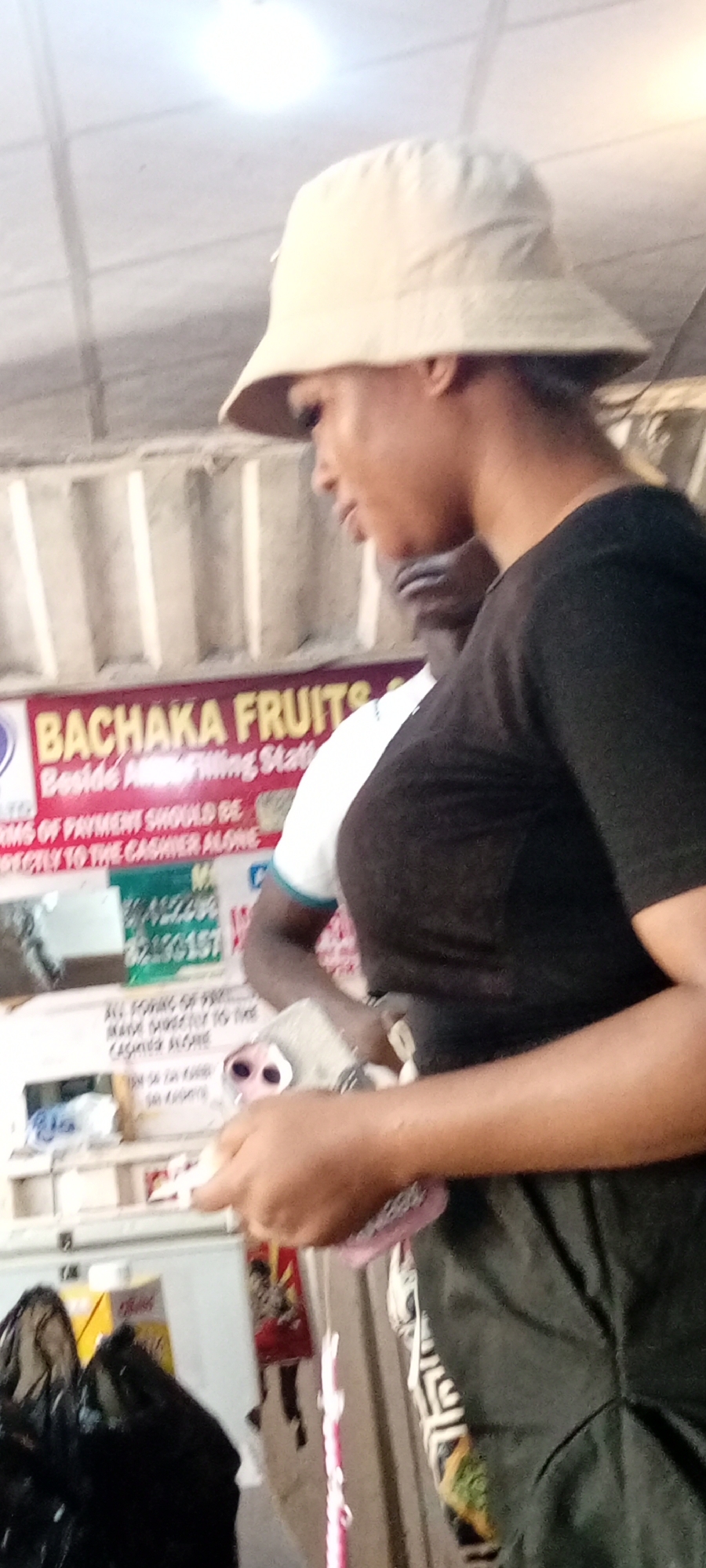 A medium shot captures a side profile of a person wearing a tan bucket hat and a black t-shirt, with a person in a white shirt visible behind them. The setting appears to be an indoor market or shop, suggested by the signs and refrigerator visible in the background. The person in the foreground is looking down and to their right, holding a pink item, possibly a phone case, in their hands, which are resting on their lap. The lighting is bright, likely from overhead fluorescent lights, creating a somewhat washed-out effect.
A prominent sign behind the individuals reads "BACHAKA FRUITS Beside Filling Stat." Another partial sign below it states "RMS OF PAYMENT SHOULD BE RECLY TO THE CASHIER ALONE." A phone number, "190157," is also visible on a sign. The overall impression is a candid moment captured in a commercial setting, possibly a fruit stand within a larger market. The exact time of day and weather are not discernible from the image. The scene is bustling with activity, indicated by the presence of multiple people and merchandise.