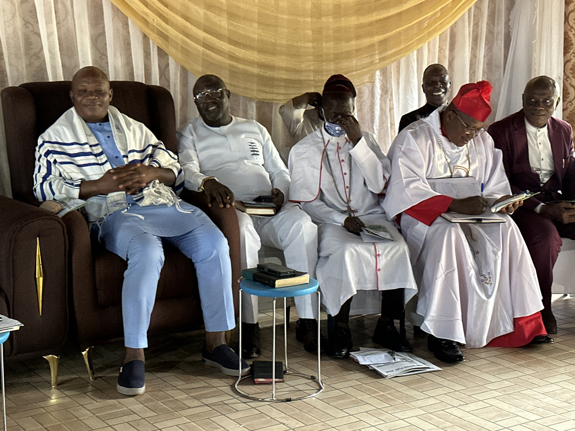 In Barden Yana, Nigeria, a group of five men are seated in chairs. The leftmost man, dressed in a blue shirt and a striped white and blue robe, sits with his hands clasped. Next to him, a man in a white robe with eyeglasses smiles and looks forward. The third man from the left wears a white robe with a red trim and a red cap, and is looking down, appearing to write in a book. He is also wearing eyeglasses. To his right, another man in a white shirt and a burgundy jacket is visible. The rightmost man, partially obscured, also appears to be wearing formal attire. They are seated in a room with a light-colored tiled floor and a backdrop of yellow and white drapes. A small blue stool with books and a phone sits in front of the third man from the left.