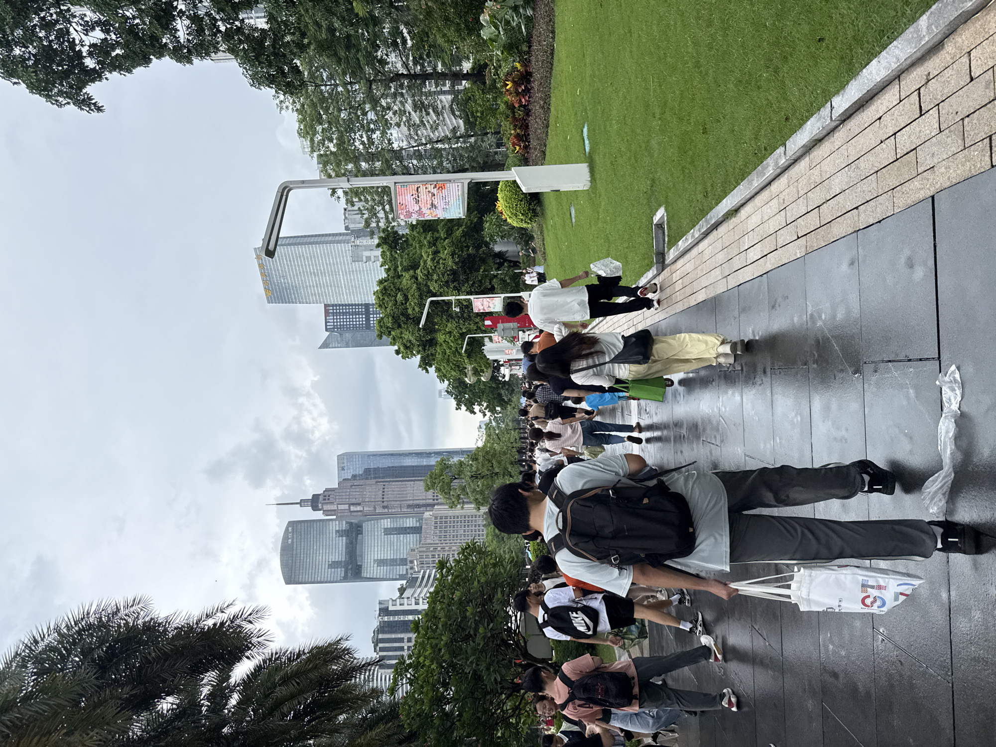 The image shows a group of people walking on a wet, paved walkway next to a well-maintained grassy area. The scene appears to be an urban park or plaza, with modern skyscrapers visible in the background. The sky is overcast, suggesting a cloudy or rainy day.
The subjects are a diverse group of individuals, many of whom are carrying bags or backpacks. Some are walking alone, while others appear to be in groups. One man in the foreground is wearing a light blue t-shirt and dark pants, with a black backpack slung over one shoulder. He is holding the handles of a white tote bag. Another person, further back, is wearing a white t-shirt and dark pants, also carrying a backpack. A person in a yellow top and white pants is walking away from the camera. The expressions on the faces of the people are not clearly visible, but their postures suggest they are moving purposefully.
In the background, several tall buildings with contemporary architecture dominate the skyline. These buildings have numerous windows and appear to be office buildings or residential complexes. Trees and some flowering plants are also visible, lining the walkway and the edge of the grassy area. Signs on poles are also present, with some featuring colorful graphics and text, though the text is not clearly legible. The ground is wet and reflective, indicating recent rain.
The overall atmosphere is that of a busy public space on an overcast day. The presence of skyscrapers and the style of the buildings suggest an urban environment, likely in a developed city. The scene depicts people going about their daily activities in this setting, possibly commuting, sightseeing, or simply enjoying the park area.