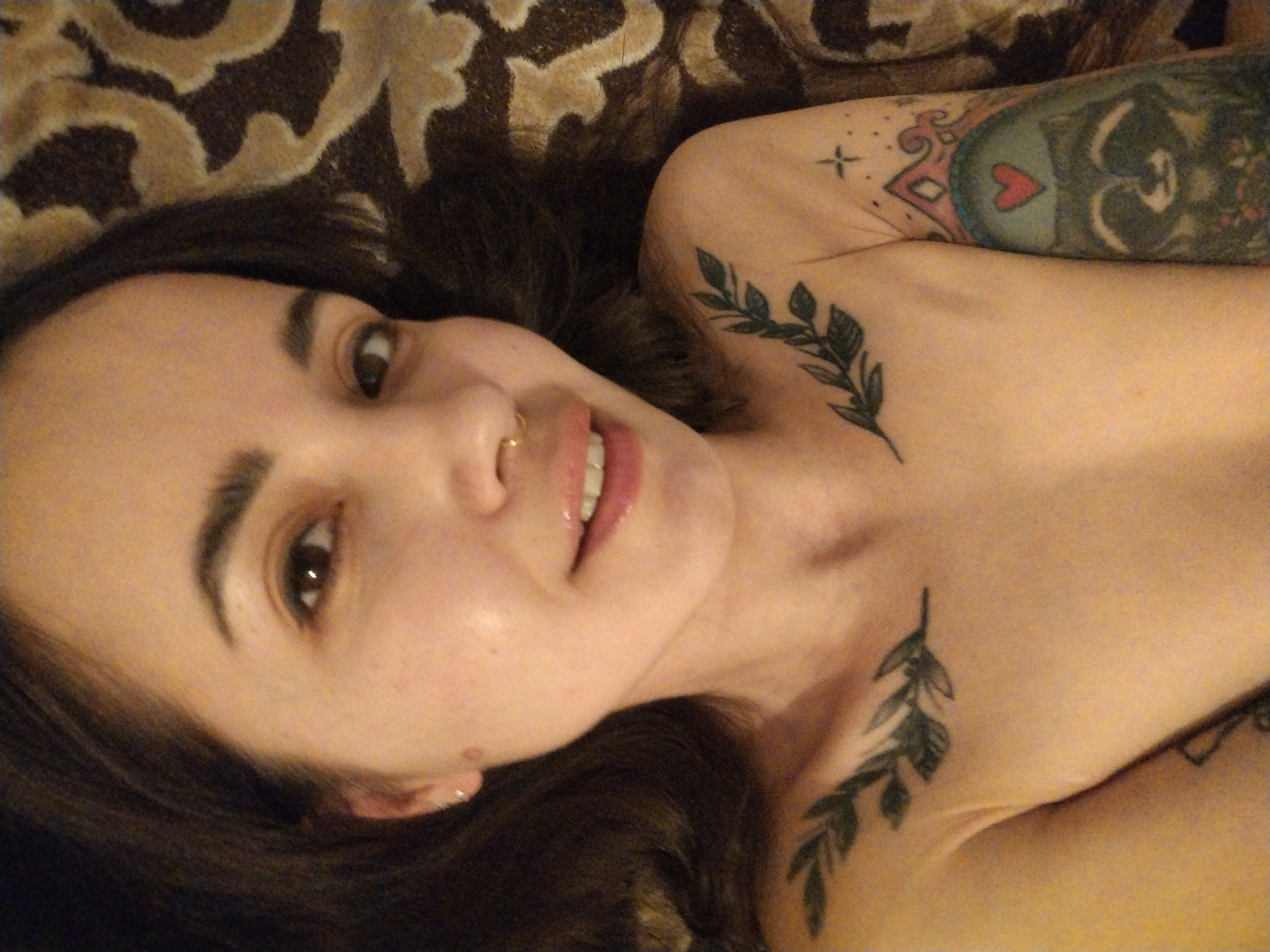 A light-skinned woman with brown hair is lying on her back on a brown and beige patterned surface. She is looking upwards at the camera with brown eyes and has a gold nose ring. She has two leaf-branch tattoos on her chest and shoulder as well as a larger tattoo on her arm that features a heart and an owl-like figure. The location is not identifiable as Noviy, Russia.