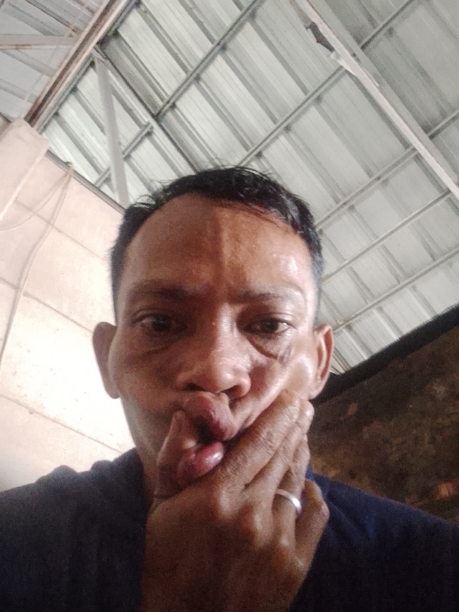 A man with short, dark hair is holding his mouth with his hand. He is wearing a dark blue shirt and a silver ring on his left ring finger. The background shows the interior of a building with a corrugated metal roof. The location is Bukit Ketok, Indonesia.