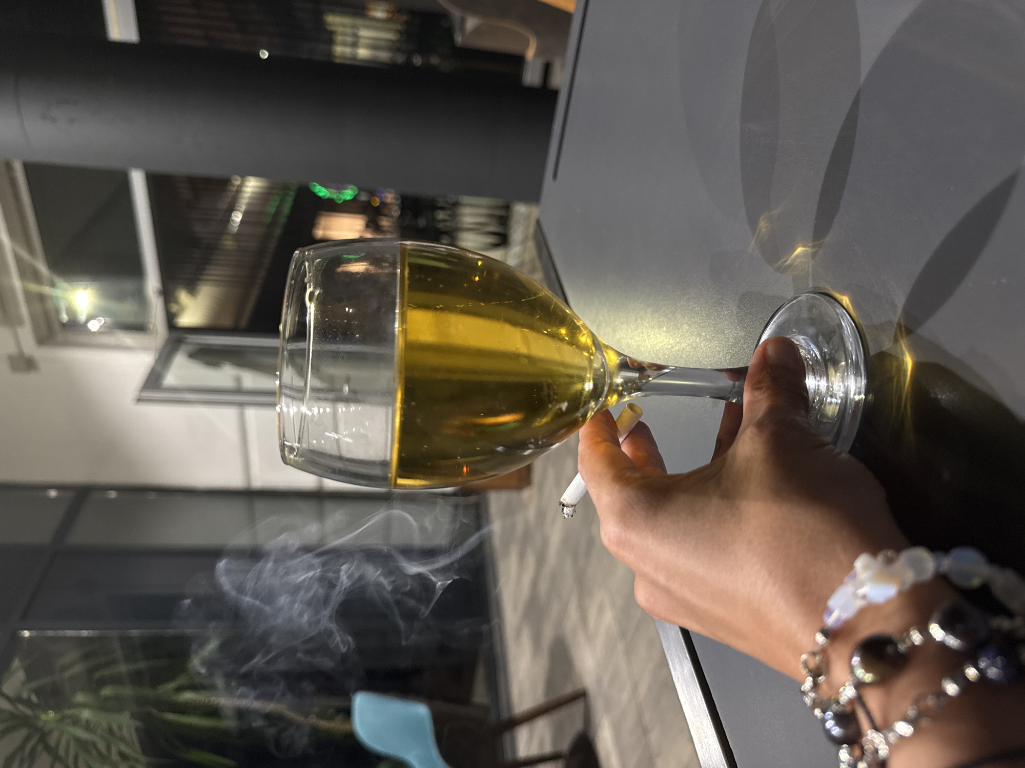 A close-up, eye-level shot shows a person's hand holding a lit cigarette and a wine glass filled with a golden-hued liquid. The hand, adorned with beaded bracelets, is positioned to the left of the wine glass. Smoke from the cigarette drifts upwards and to the left. The wine glass is on a dark gray table. In the background, a plant is visible to the left, and a large black pillar is on the right. The setting appears to be an indoor establishment, possibly a restaurant or bar, with windows in the background showing a dimly lit urban scene, suggesting the location is Ilidža, Bosnia and Herzegovina. The overall lighting is dim, with reflections from the wine and ambient light creating highlights.
