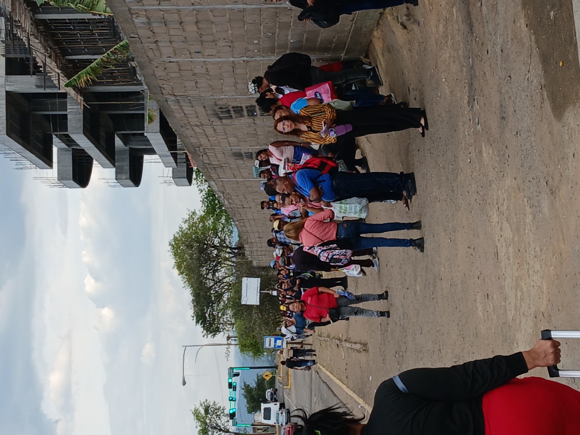A daytime shot depicts a crowd of people lining up along a sidewalk in Ciudad Guayana, Venezuela. They stand along a concrete wall beside an unfinished, multi-story building. The people are of mixed ages, wearing casual clothing, with some carrying bags. Behind them, a street with traffic lights and vehicles, including cars and a van, is visible. The weather appears clear, with a cloudy sky. A person in the foreground is holding the handle of a wheeled suitcase.