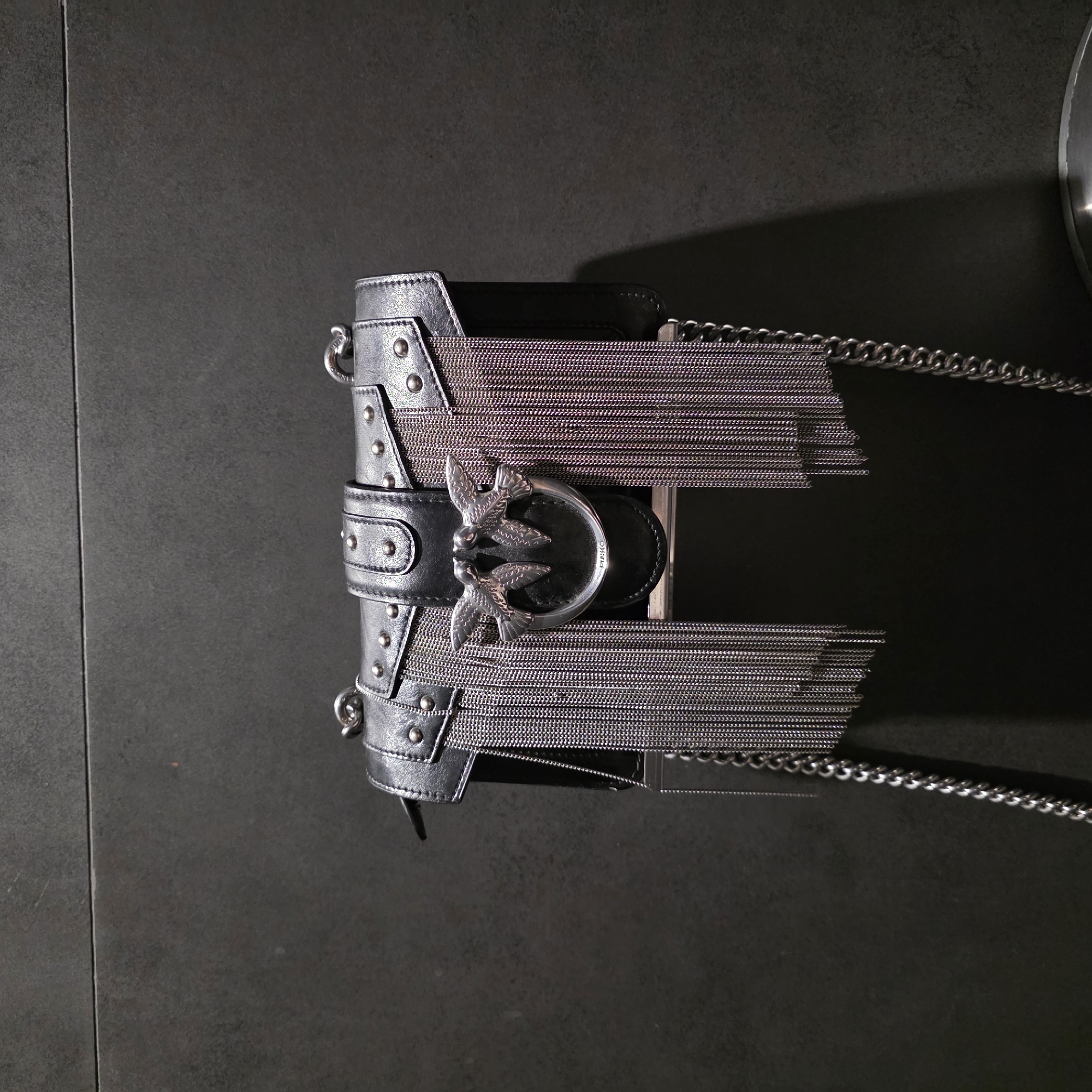 An overhead shot displays a black leather handbag or clutch placed on a dark, textured surface, possibly a tiled floor or tabletop. The bag is oriented vertically in the image but appears to be designed to be worn horizontally.
The handbag is crafted from black leather, possibly faux leather, and adorned with numerous silver-toned metal details. Small, round studs punctuate the edges and a central horizontal strap. The most prominent feature is the unique clasp, a circular silver-colored metal buckle, intricately designed with two stylized birds, likely swallows or doves, facing each other with their wings spread. The brand name "PINKO" is clearly engraved on the clasp.
Cascading from either side of the central clasp are decorative fringes made of multiple thin, silvery chains, adding a shimmering, layered effect to the bag's design. A thicker, silver link chain strap is also visible, extending from the top right of the bag, indicating it is likely a shoulder or crossbody style.
The background is uniformly dark gray with a subtle texture, providing a stark contrast that makes the metallic details and the bag stand out. Shadows cast by the bag suggest direct, artificial lighting. There are no people or discernible activities, and no specific location cues are visible within the image beyond the minimalist, indoor-like setting.