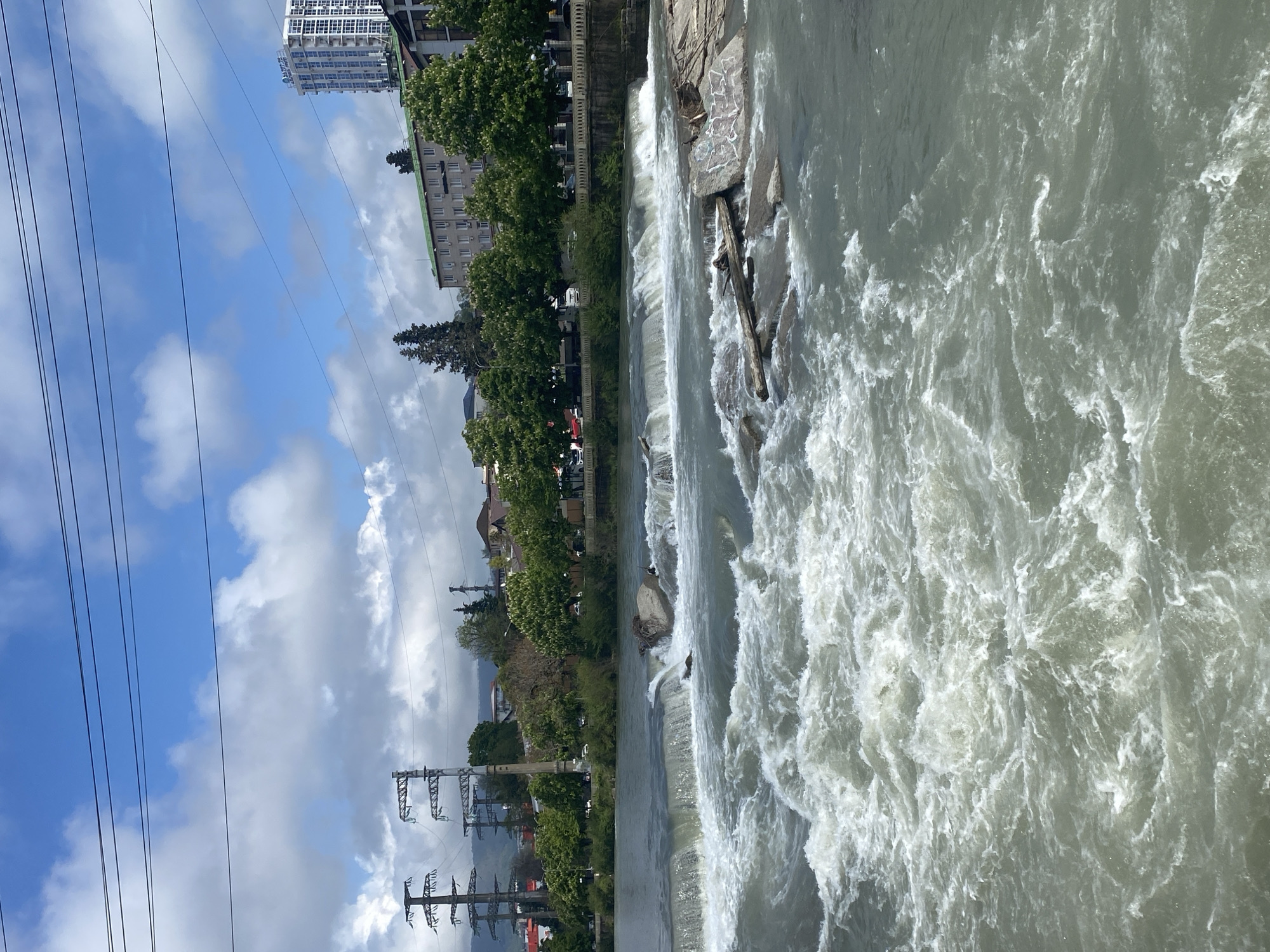 The image shows a river scene in the city of Sochi, Russia. The river flows with white water and rapids, cascading over a series of small dams or weirs. Along the riverbank are buildings, trees, and power lines. The sky is partly cloudy with a mix of blue sky and white clouds.