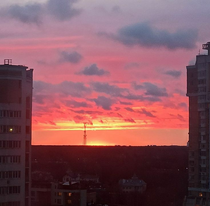 The image captures a sunset in Minsk, Belarus, framed by two tall buildings. The sky is filled with pink and orange hues, with clouds scattered throughout. In the distance, a tall communication tower is visible against the setting sun. Below the horizon, a dark landscape of trees and buildings is present. The buildings on either side of the frame appear to be residential, with multiple windows visible.