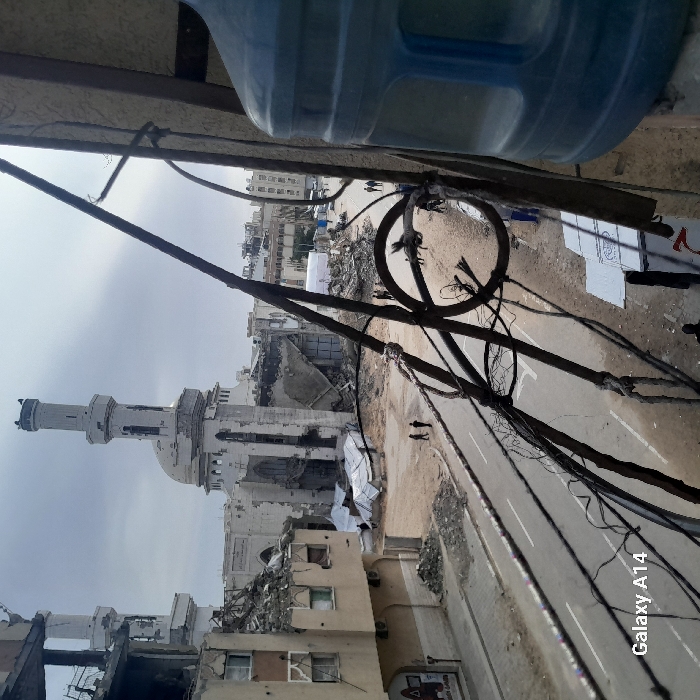 The image displays an elevated view of a severely damaged urban landscape in Khan Yunis, Palestinian Territory. In the immediate foreground, a fragment of a blue plastic water bottle is visible on the right, partially obscuring a vertical support structure. Multiple black electrical wires, some bundled and others loose, traverse the scene horizontally from the left mid-frame, attached to a decorative metal railing or support.
Below, a paved street with a faded double white line marker is visible, flanked by extensive areas of debris and rubble. Several individuals are observed walking on the street and within the rubble-strewn spaces. Two people walk on the left side of the street, moving away from the viewer. Scattered groups of individuals are also seen further down the street. White tarpaulins are spread across various ground areas, covering debris or forming makeshift coverings.
In the mid-ground and background, the Great Mosque of Khan Yunis is centrally prominent, exhibiting significant structural damage to its exterior walls and exposing internal elements, though its tall minaret and dome remain partially intact. To the left of the mosque, a multi-story building is heavily damaged, with portions collapsed and rebar exposed. Further into the background, additional damaged buildings and several white temporary structures (potentially tents or covered areas) are visible along the street. The sky is overcast and uniformly grey. A "Galaxy A14" watermark is present in the lower-left corner of the image.