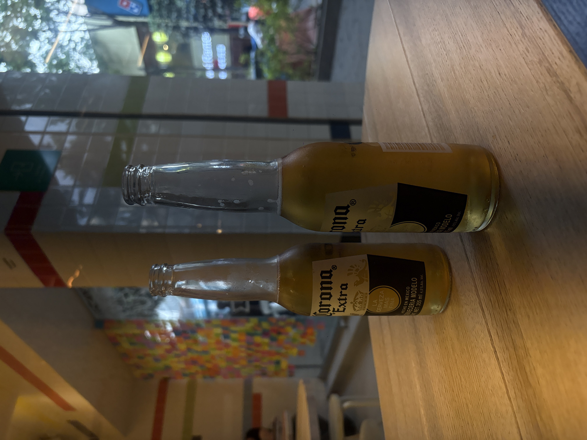 The image shows two Corona Extra beer bottles lying on a wooden surface. The bottles appear to be partially empty. In the background, a window reflects a cityscape, suggesting the bottles are indoors. The window also shows a building with colorful art. The bottles are labeled in Spanish, stating they are made in Mexico by Cerveceria Modelo. The text provides information about the alcohol content and the volume of the bottles.
