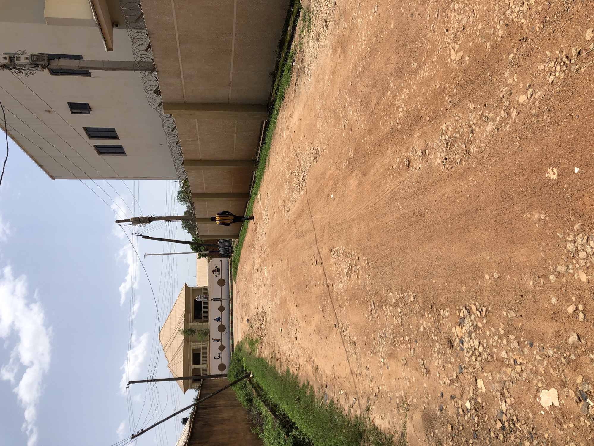 A person wearing a yellow and black vest is walking away from the camera down a dirt road in Bar Jirgi Summa, Nigeria. The road is unpaved and appears to be in disrepair, with gravel and rocks scattered across its surface. On the right side of the road, there is a light-colored, concrete wall topped with barbed wire. Behind the wall, a portion of a white building with multiple windows is visible. On the left side of the road, there is a brown wall, and beyond it, lush green vegetation. In the distance, at the end of the road, a light-colored building with a beige roof is visible, with a white decorative wall in front of it featuring a mural of children. Utility poles and wires are present throughout the scene, indicating the presence of infrastructure. The sky is partly cloudy.