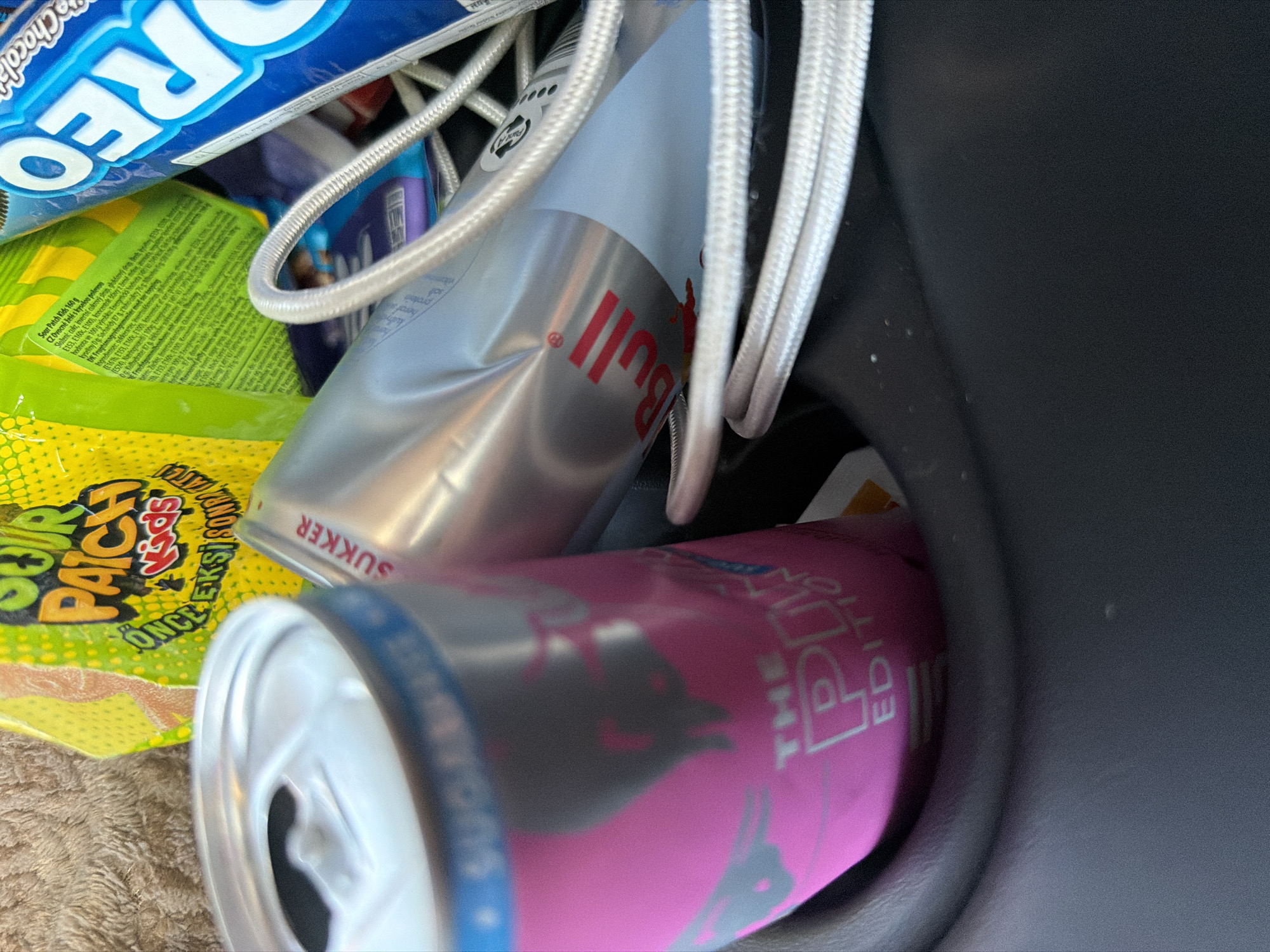 The image shows a close-up view of a car's cup holder containing several items. In the foreground, there is a pink can with the text "THE POP EDITION" visible. To the right of the pink can, a silver can with "SUKKER" and the Red Bull logo is partially visible. In the background, there are bags of candy, including "Sour Patch Kids" and a blue package of Oreo cookies. A white braided cord is also present, wrapped around some of the items. The background appears to be a textured surface, possibly a car seat or dashboard. The lighting suggests an indoor or shaded environment. There are no people visible in the image, and no specific location in Gothenburg, Sweden can be identified from the contents.