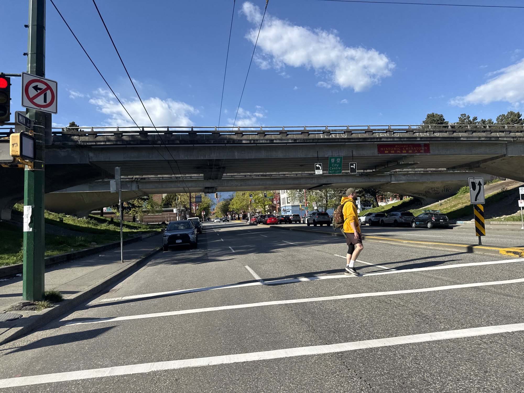 The image depicts a street scene in Vancouver, Canada, specifically in the Historic Chinatown area. A man wearing a yellow hoodie, brown shorts, black knee brace, and a backpack is crossing the street within a crosswalk. Above the street, an overpass bears a sign that reads "Welcome to Historic Chinatown" in English and Chinese. Signs indicating exits for "City Centre" are visible as well. Several cars are parked or driving on the street, and the sky is blue with scattered clouds. A traffic light is visible, showing a red light with a "no left turn" sign.