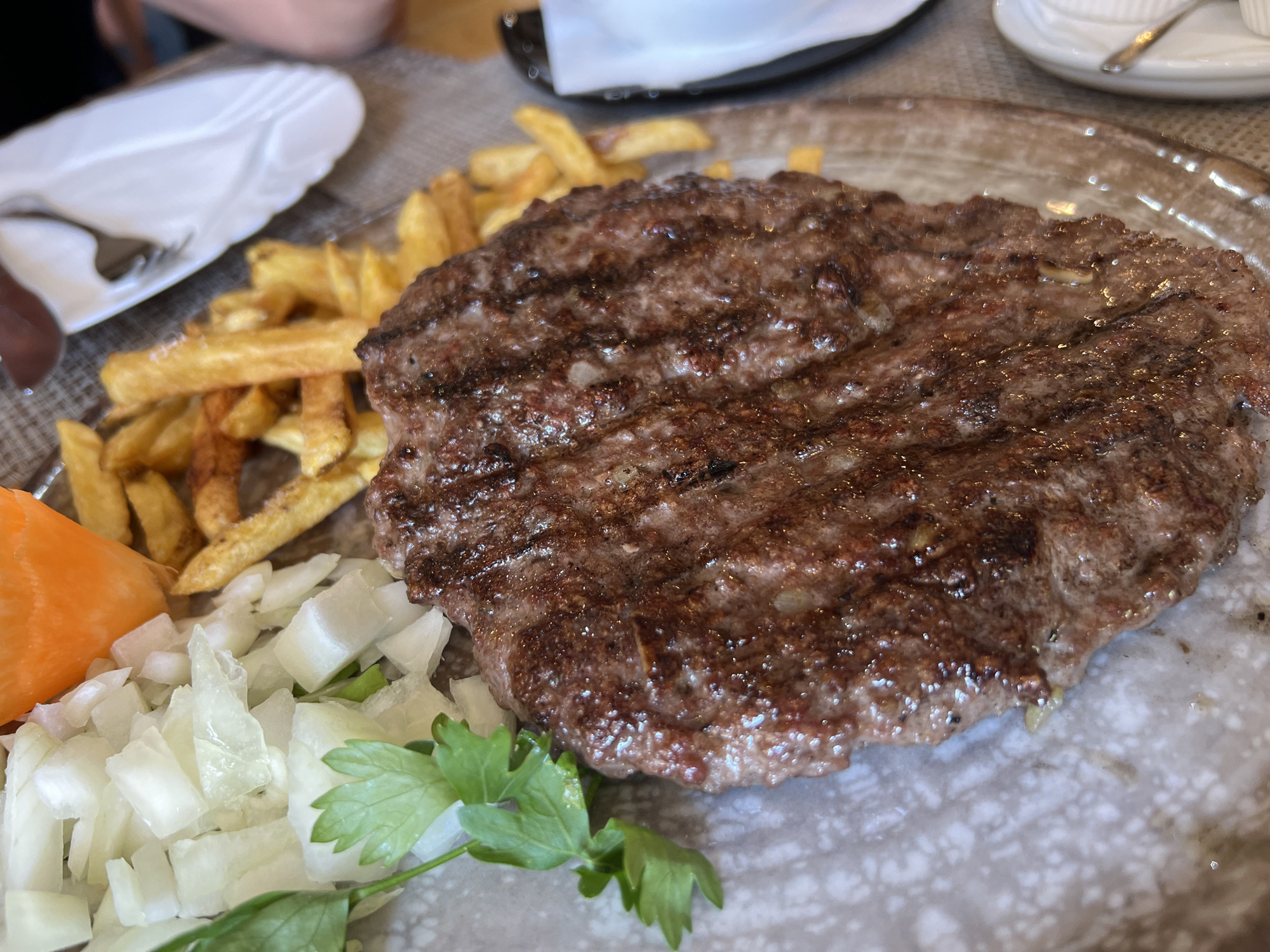 A close-up shot captures a grilled meat patty, likely a burger or ćevapi, served on a rustic plate. The patty is well-cooked with visible grill marks and glistening juices. Alongside it are golden french fries, a pile of chopped white onions, a slice of orange bell pepper, and a sprig of parsley garnish. The meal appears to be set on a table in a restaurant, with blurred background elements suggesting other diners and restaurant tableware. This meal was photographed at Budva, Montenegro, at a restaurant known for its Balkan cuisine.